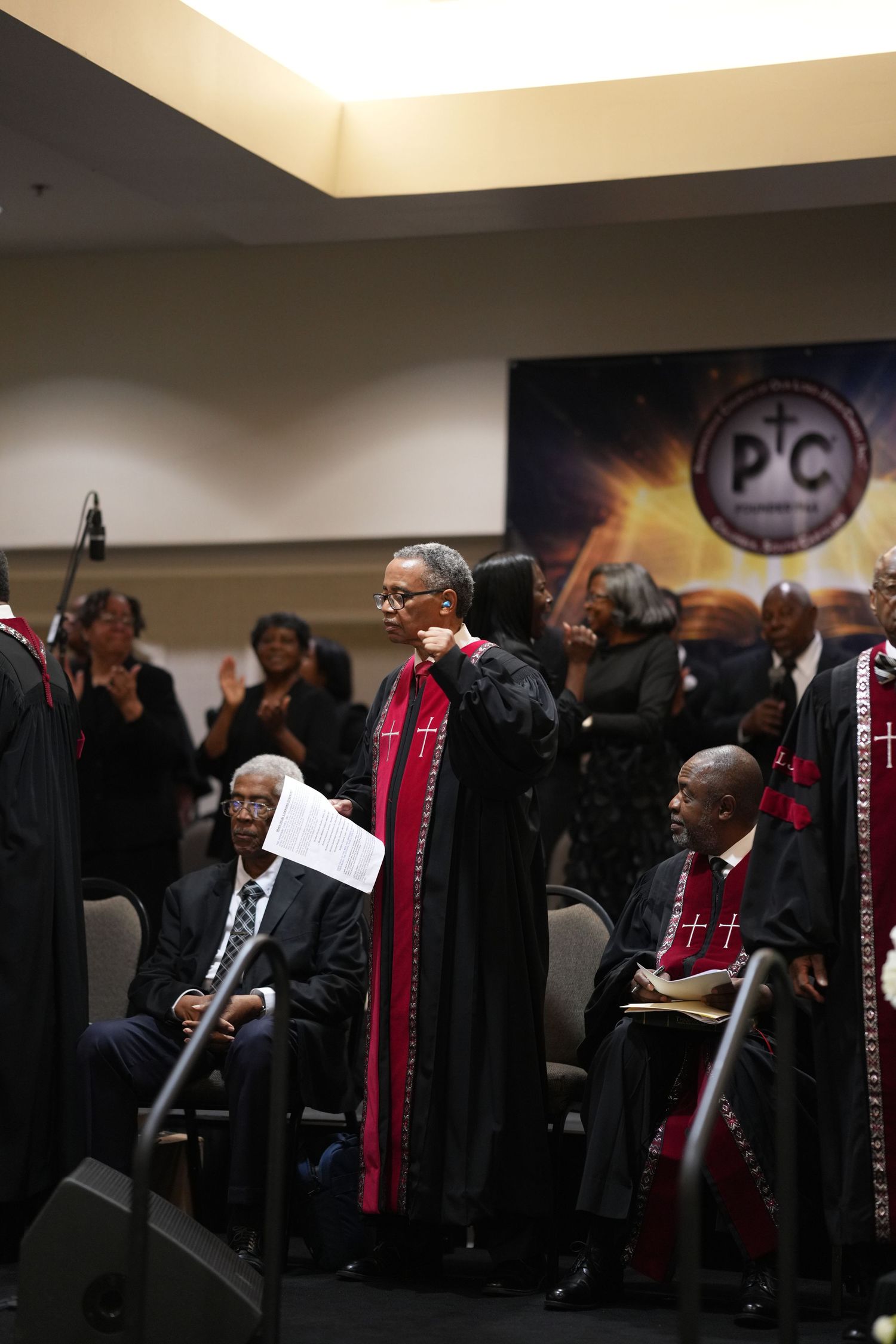 Church clergy in black and red robes stand during a formal religious ceremony in a conference hall.