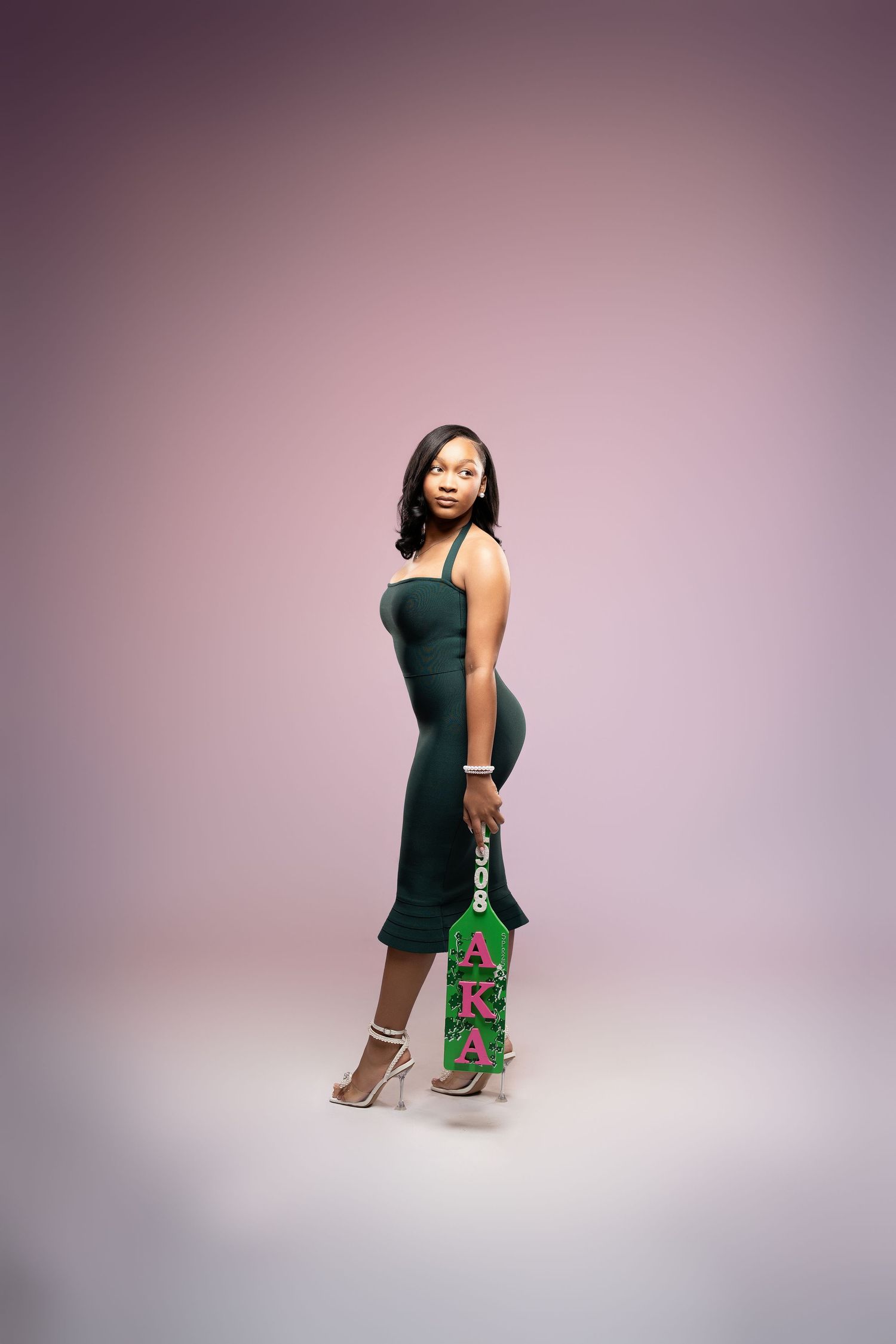 A woman in a fitted dark green dress poses elegantly with a festive holiday gift bag against a purple backdrop.
