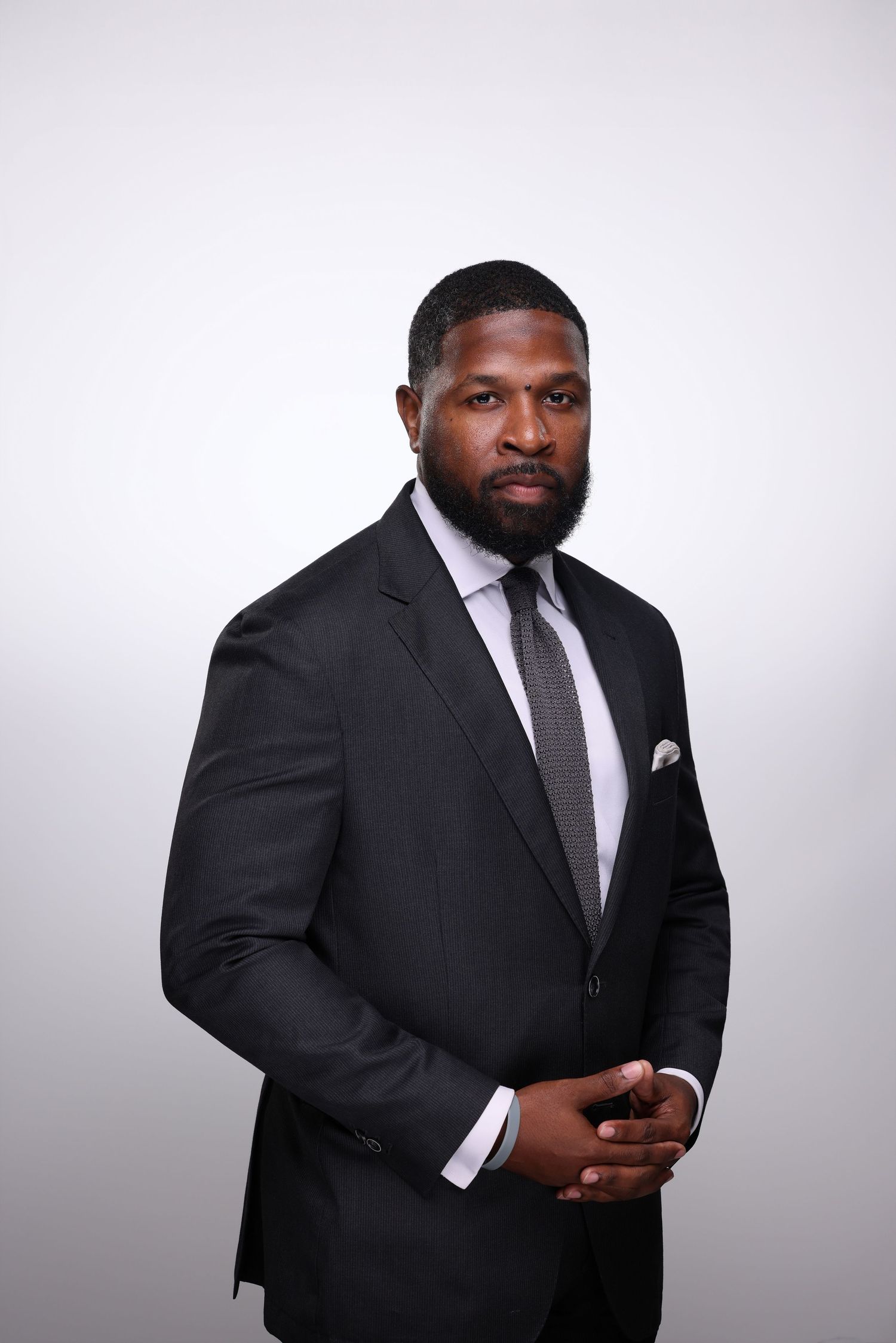A professional man in a black suit with gray tie and white pocket square poses confidently against a light gray background.
