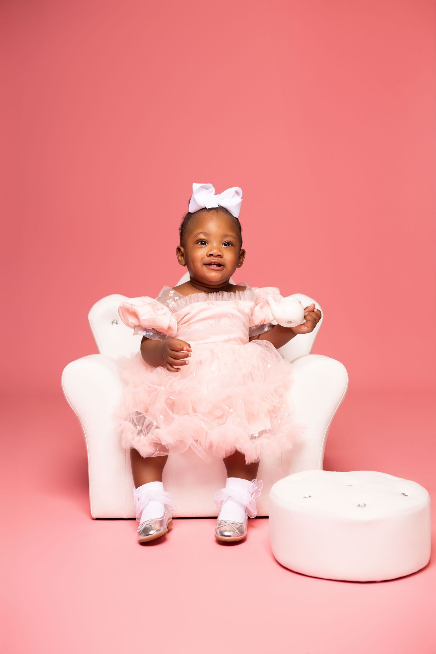 Young child in a pink tulle dress sitting on white furniture against a pink background.