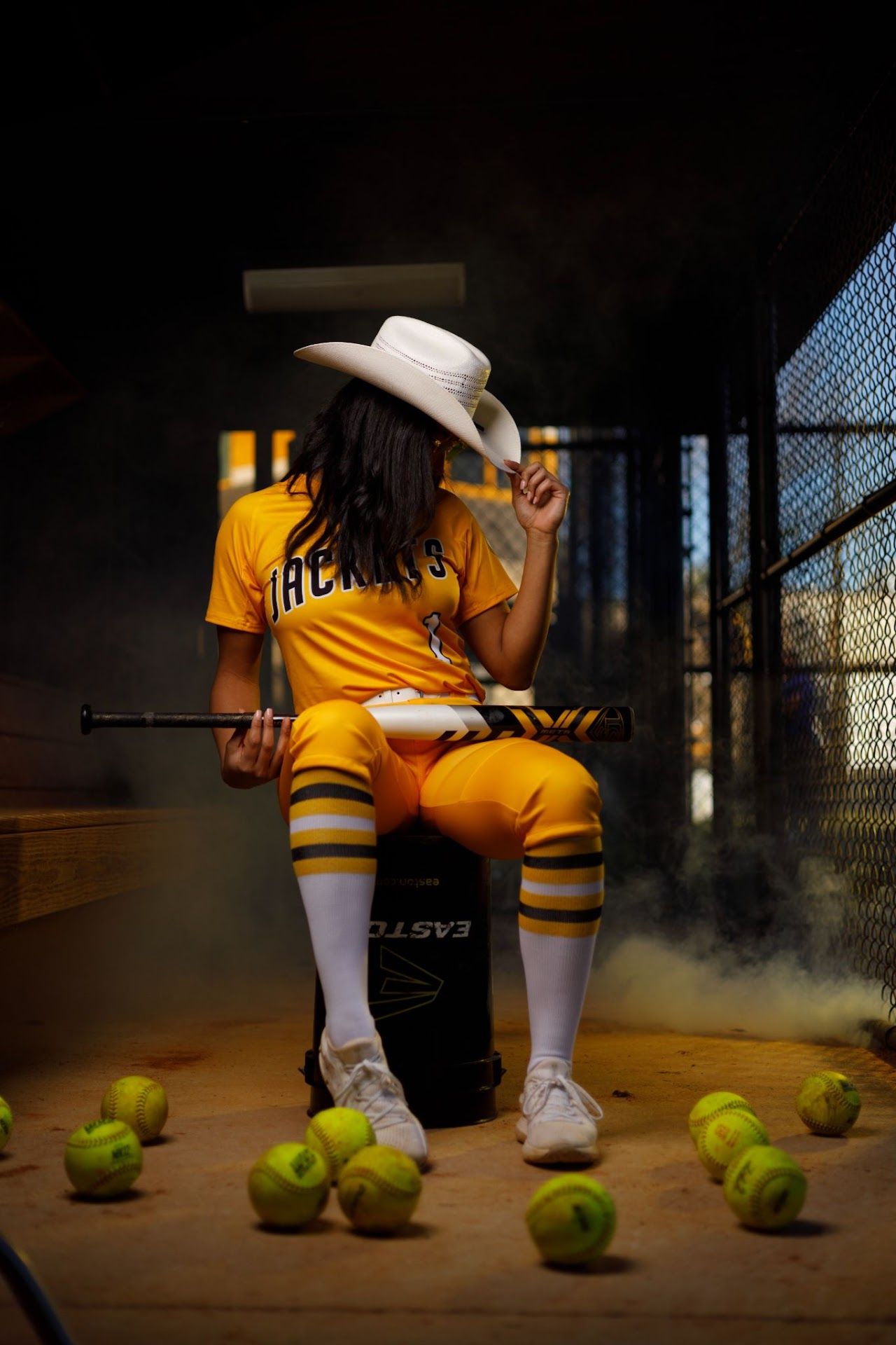 Irmo HS Softball player in yellow uniform sits in dugout surrounded by tennis balls in dramatic lighting.