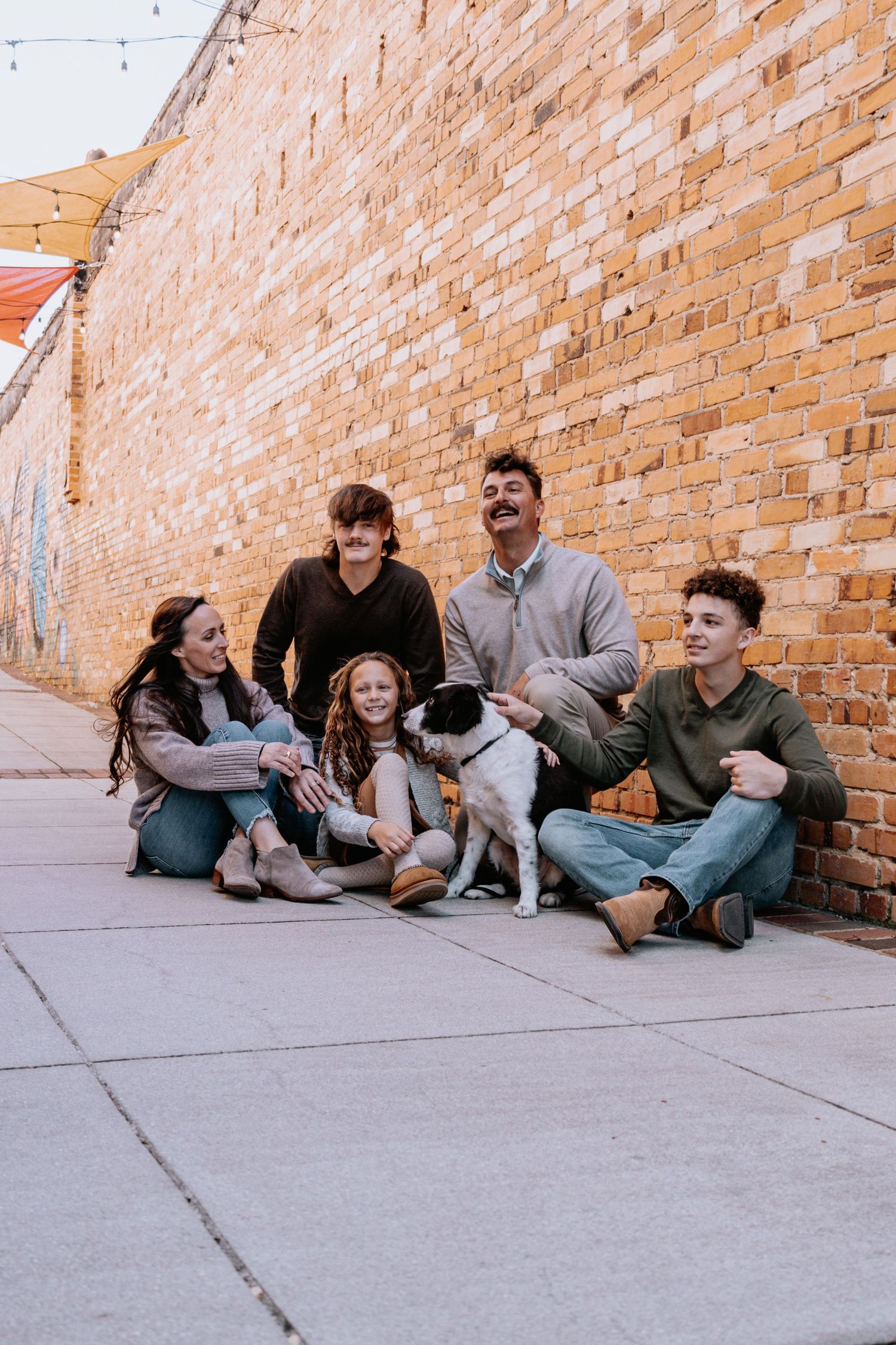 Casual group photo of people and a dog sitting against a brick wall on a sunny day.