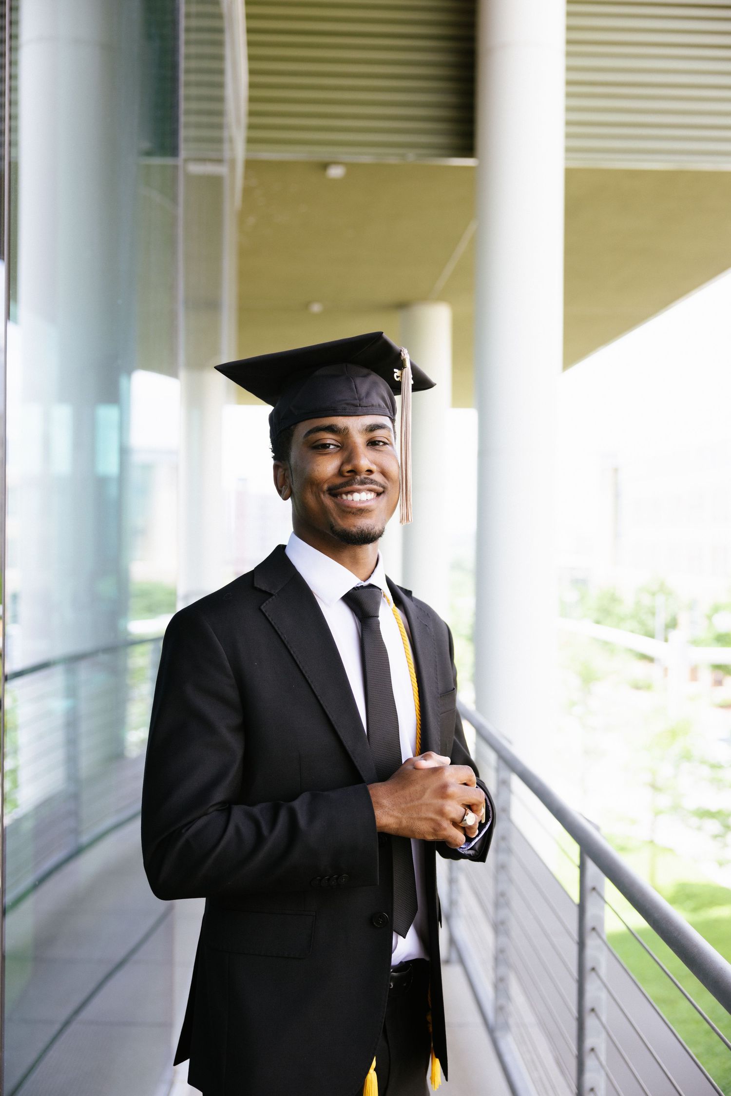 Graduate in cap and gown smiles while standing in modern building hallway.
