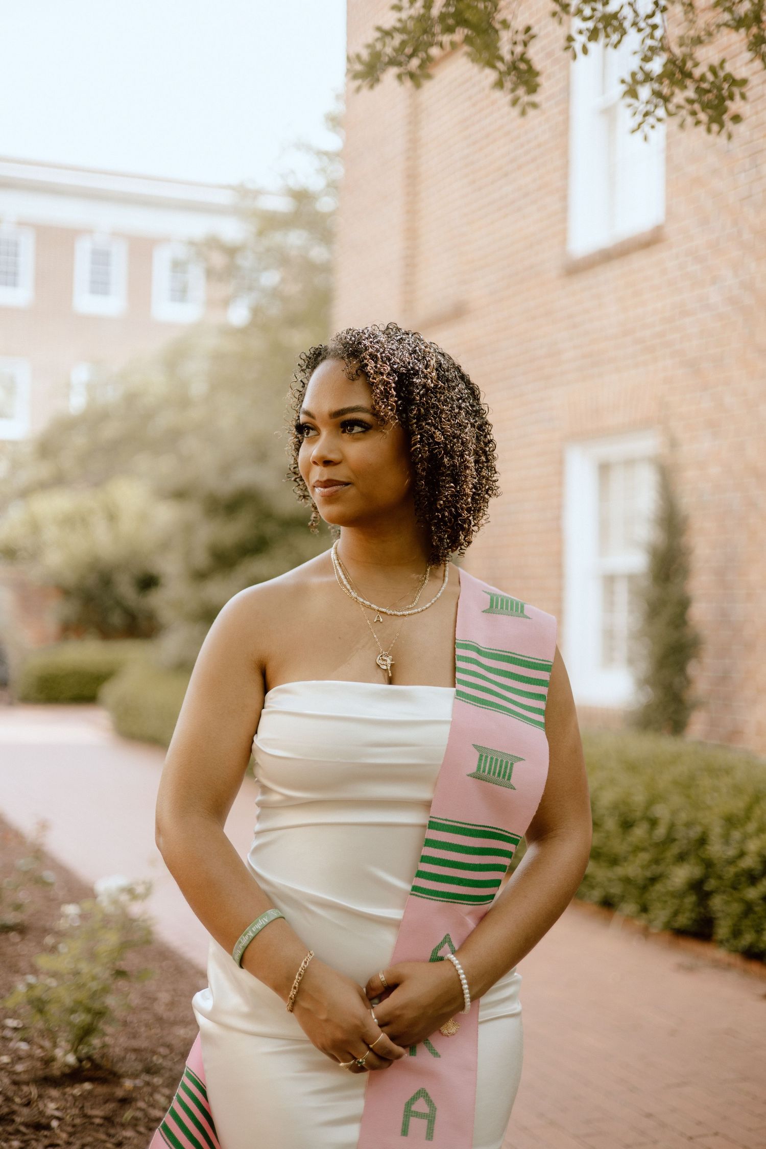 Graduate in white dress poses outdoors with traditional graduation stole.