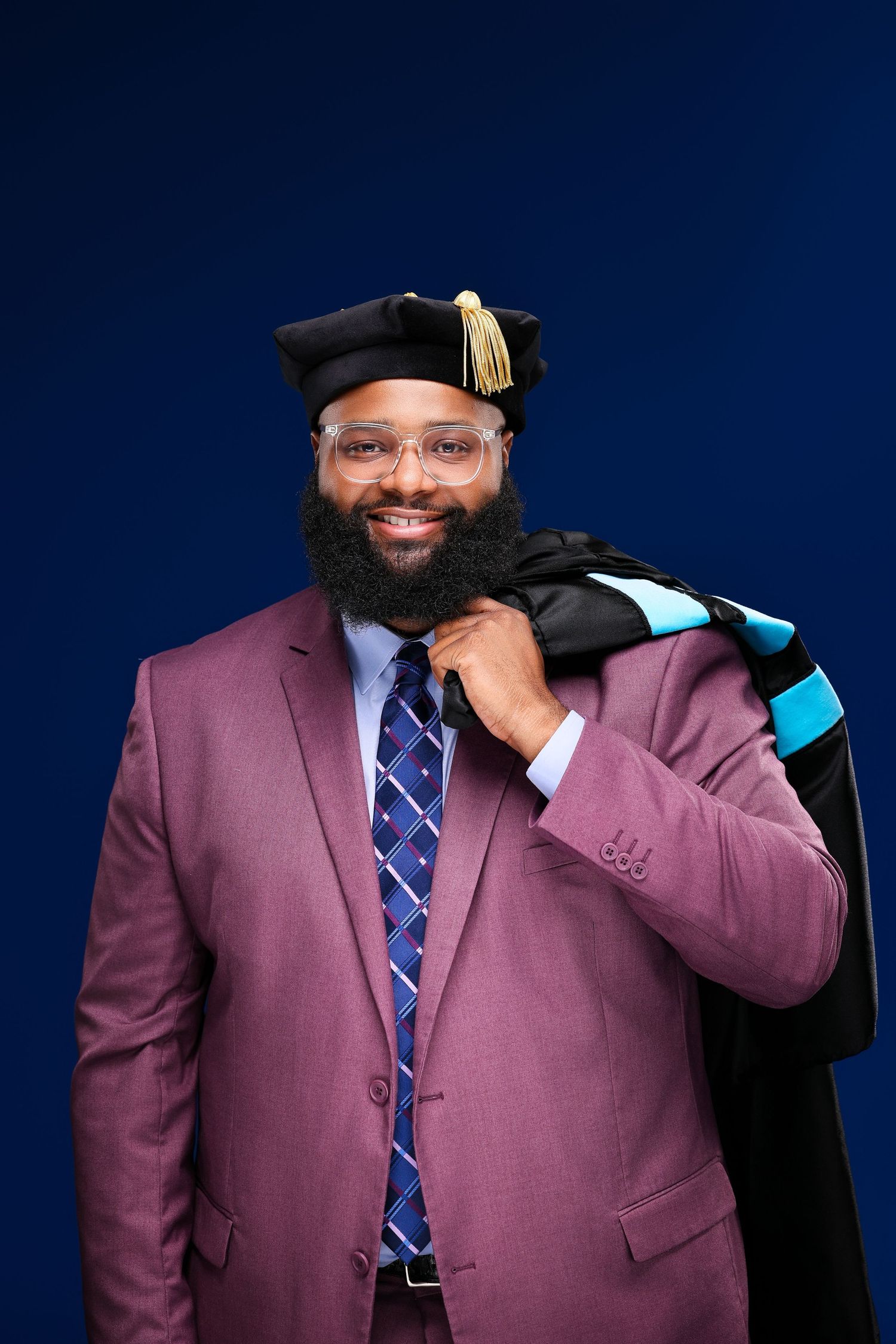 A graduate in a mauve suit and blue patterned tie poses with academic regalia against a dark blue background.