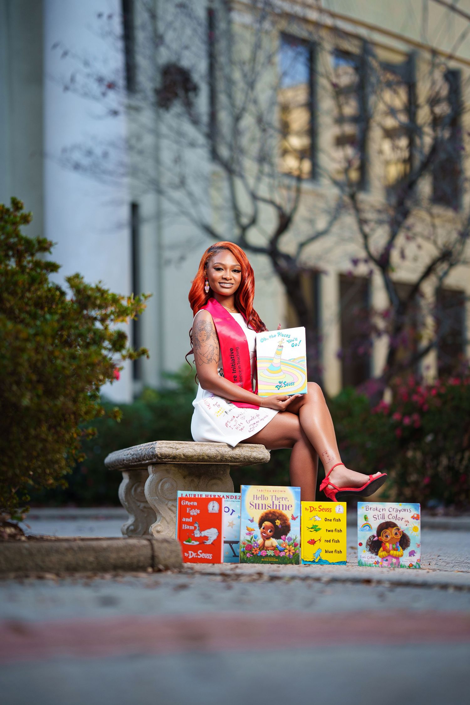 A person with vibrant red hair sits on a stone bench surrounded by colorful children's books in an outdoor setting.
