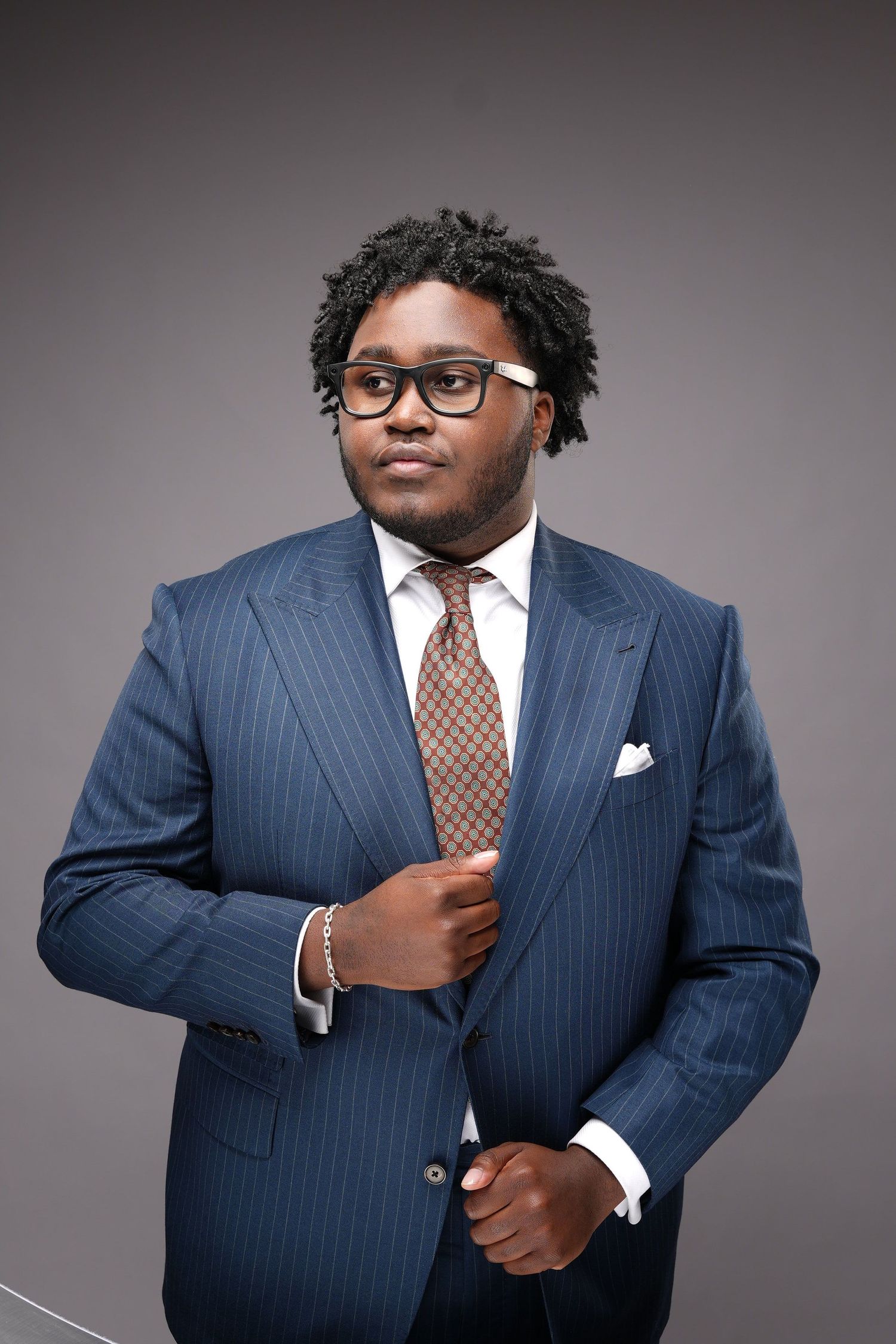 Professional studio portrait of person in navy pinstripe suit with brown tie and glasses against gray background.