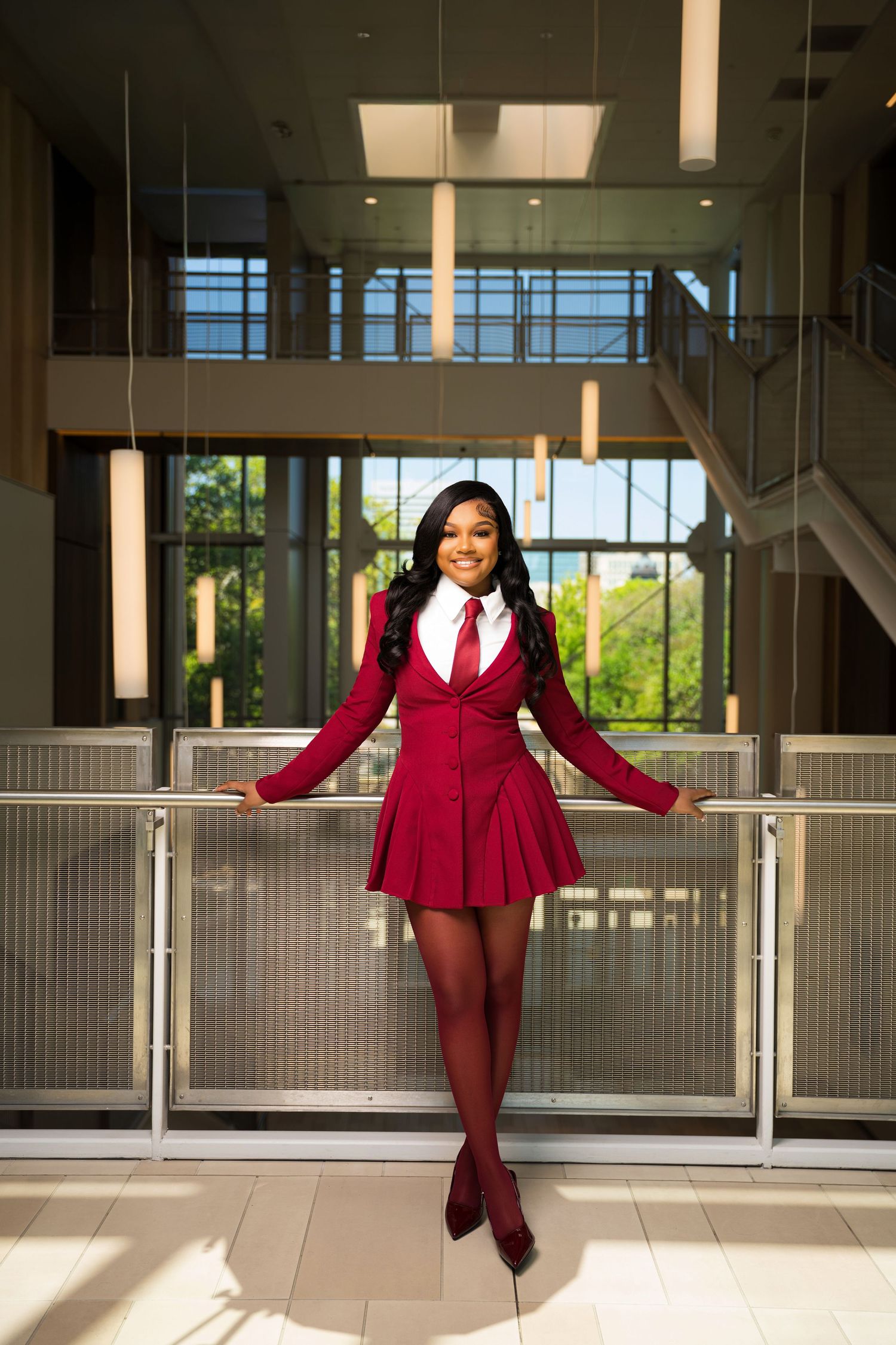A young woman poses confidently in a red blazer dress, white shirt, and tie with matching tights in a modern building.