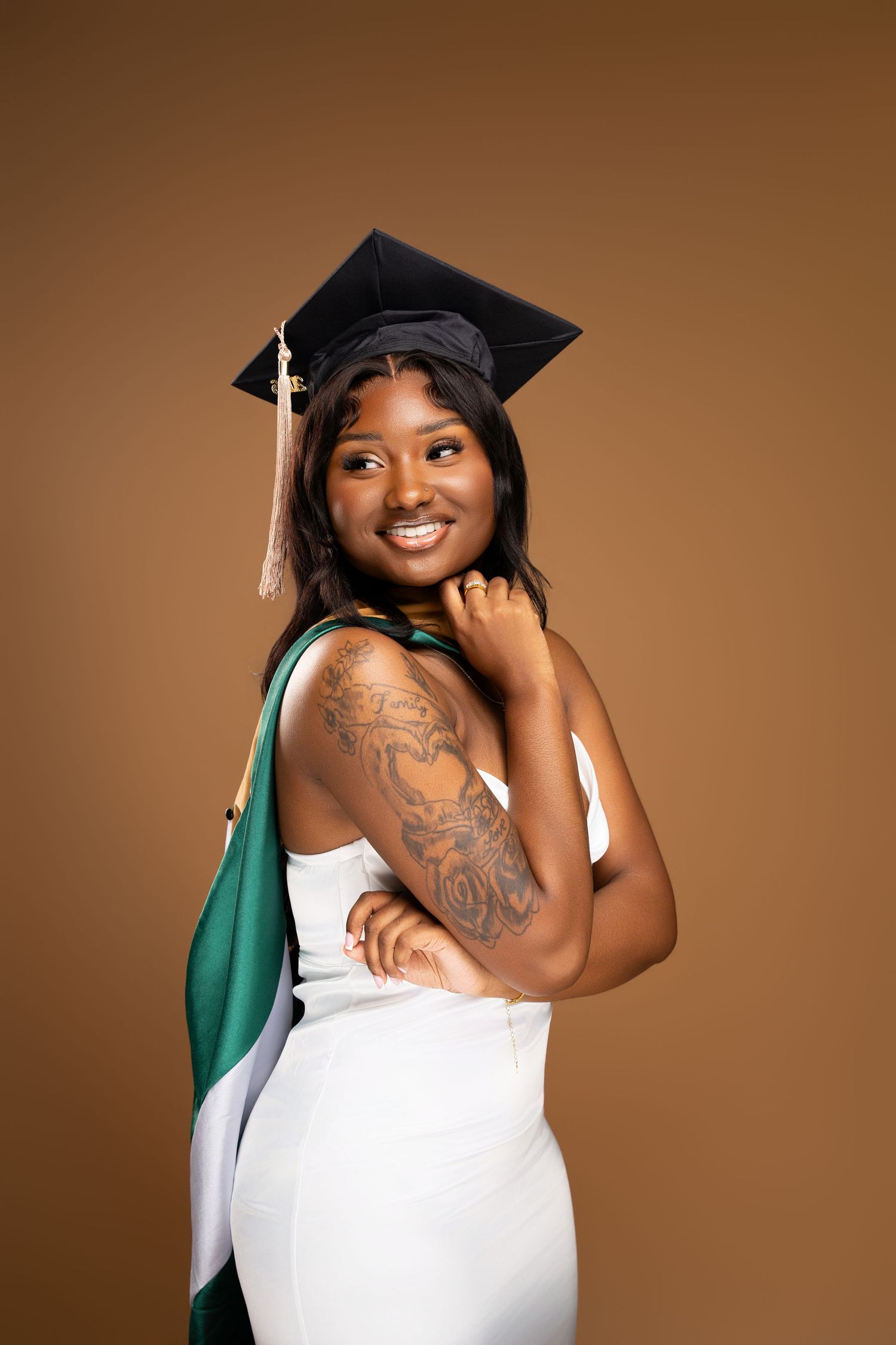 Smiling Black female graduate in white dress and black cap with green sash, showing arm tattoo, against brown background.