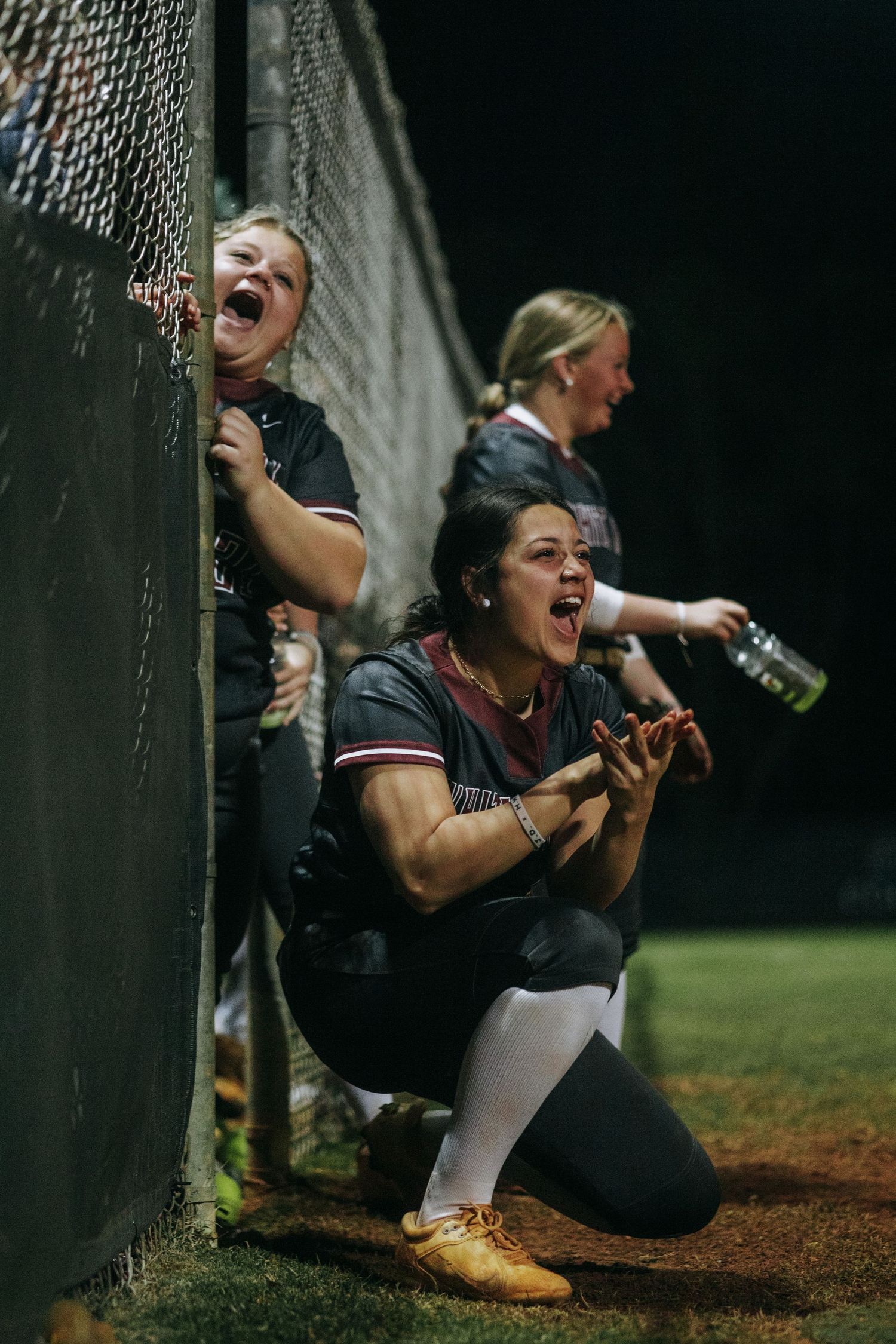 Softball players cheer enthusiastically from behind the dugout fence during a game.