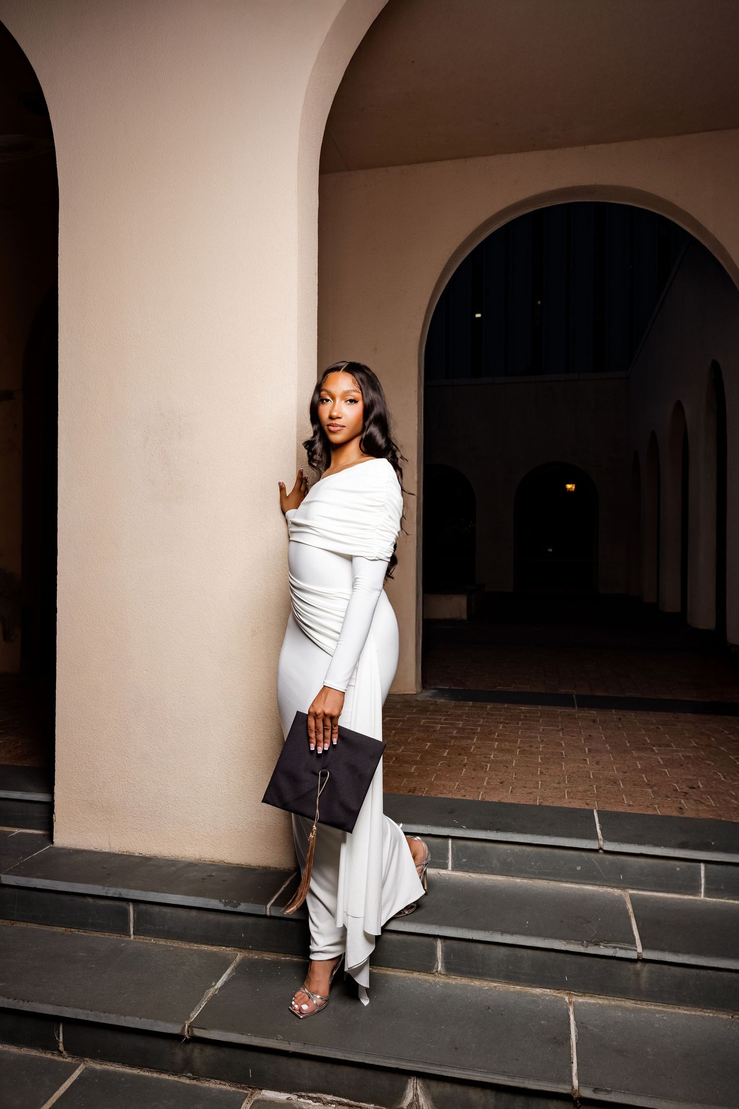 Graduate in flowing white dress holds clutch while posing on architectural steps.