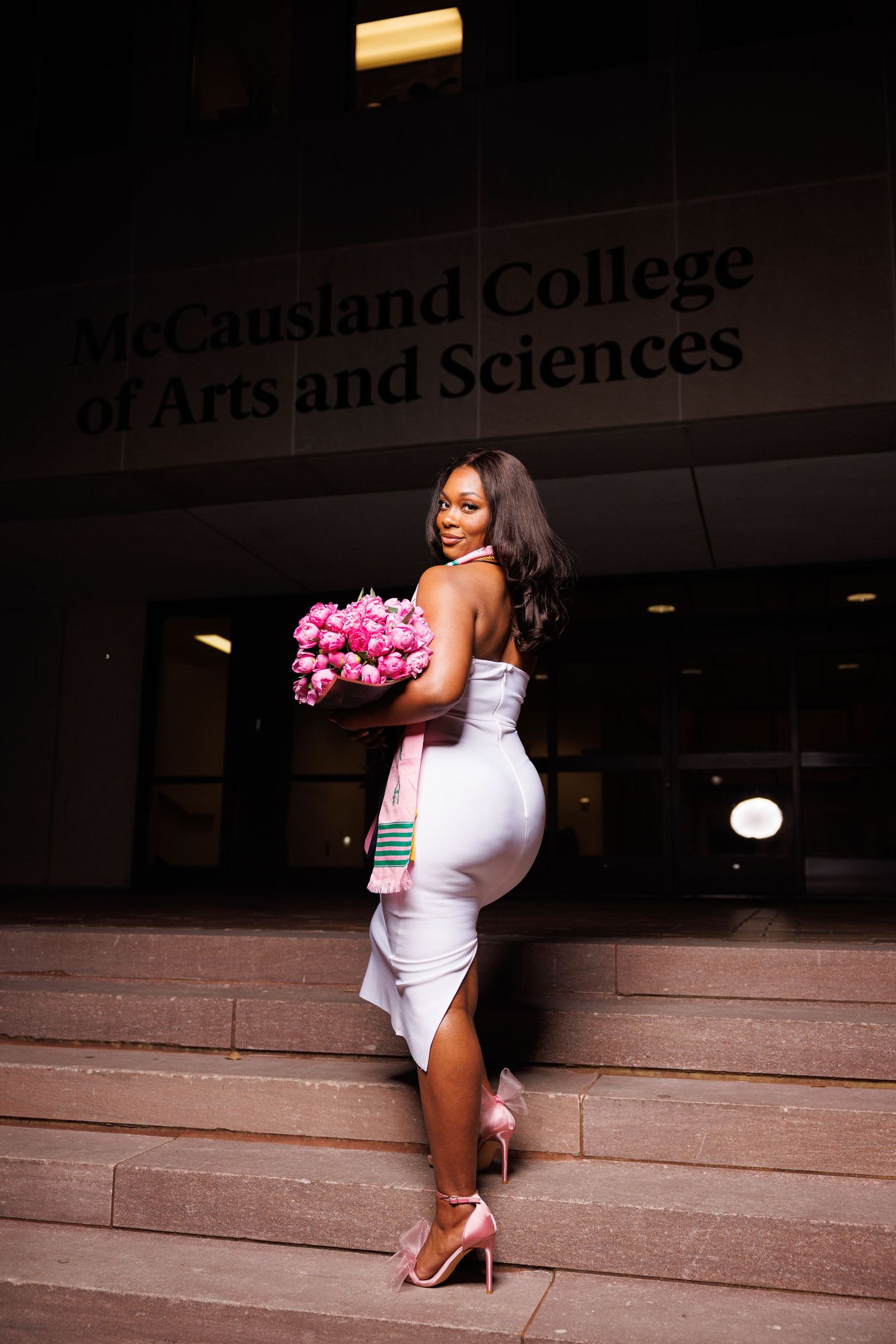 Graduate in white dress holding pink flowers on stone steps.