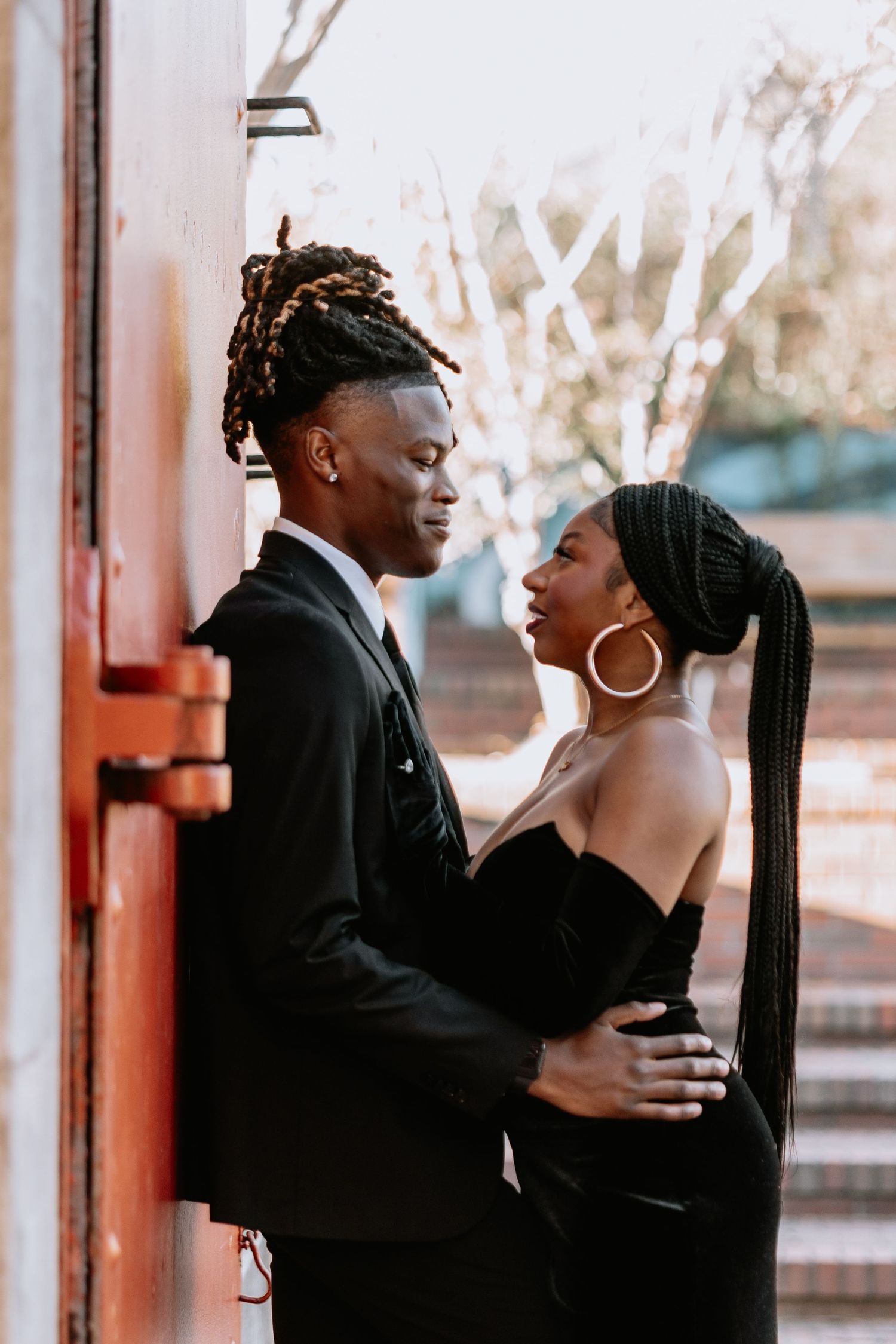 Elegant couple in black formal wear posing together against urban backdrop.