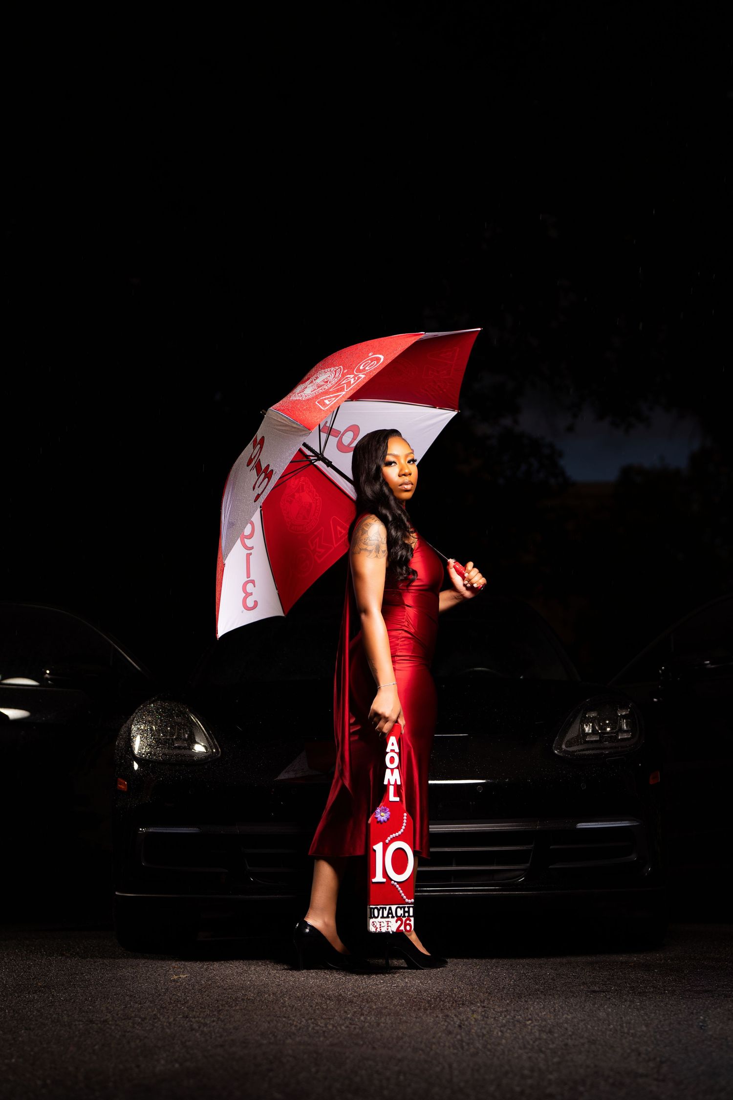 Woman in red gown holding red and white umbrella beside black car at night.