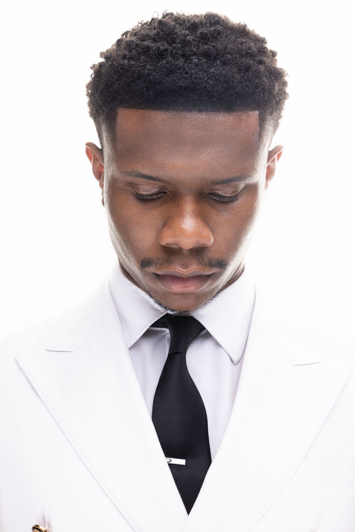 A person in a crisp white suit jacket and black tie looks down thoughtfully in a professional headshot series.
