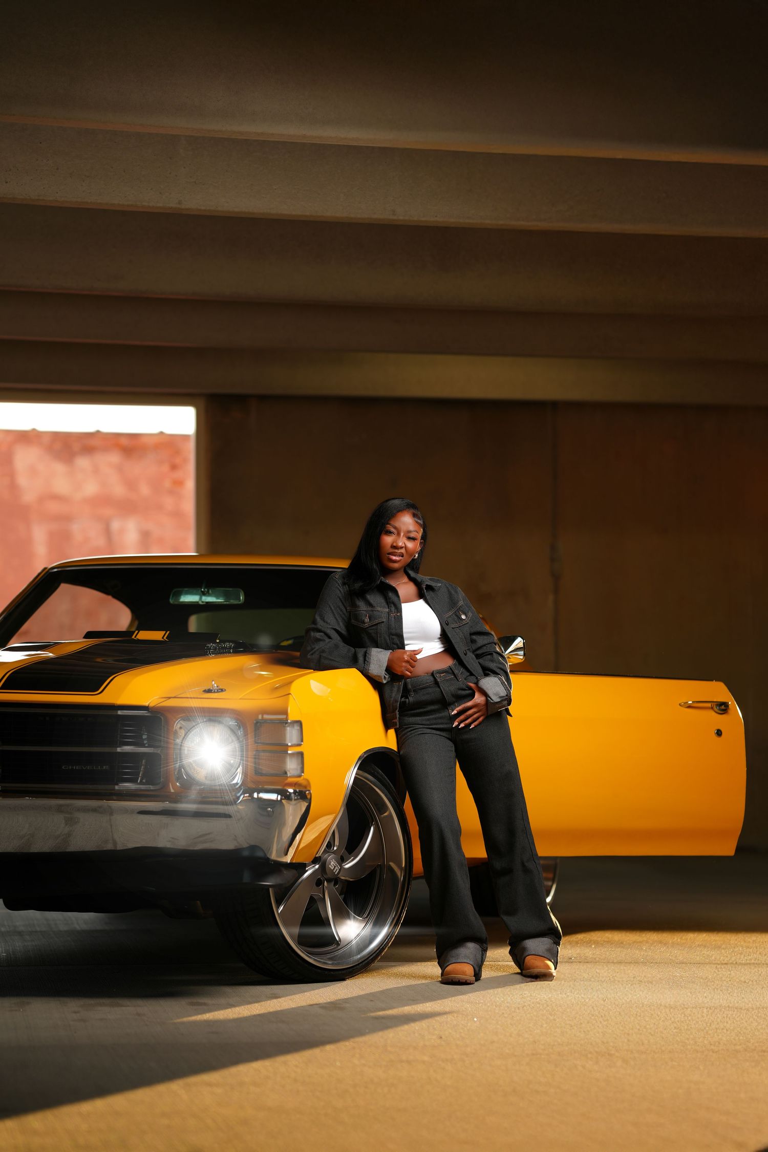 A young Black woman in a denim jacket leans against a yellow classic muscle car in a dimly lit parking garage.