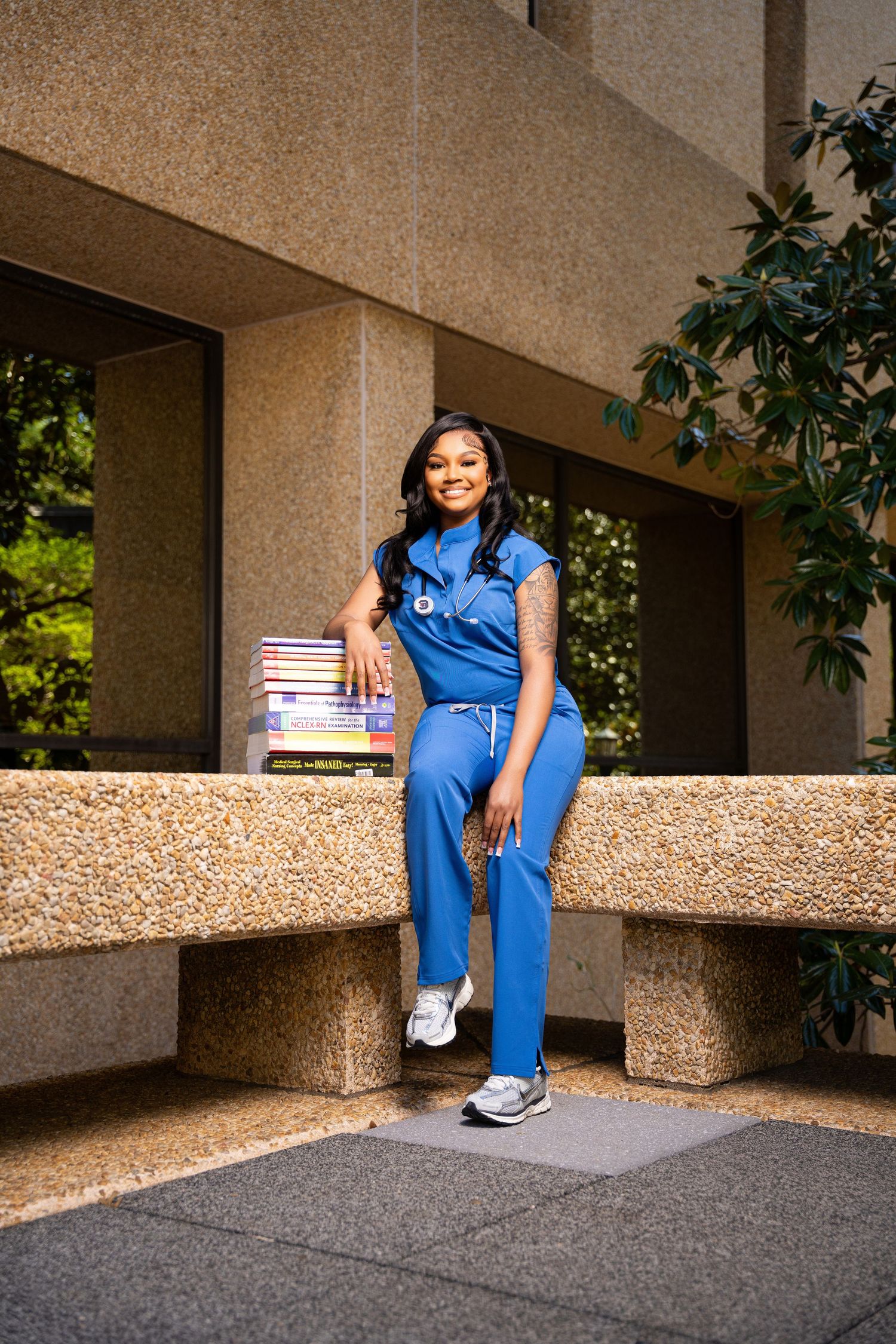 Young nurse in blue scrubs sitting outdoors holding textbooks, stethoscope around neck, smiling confidently.