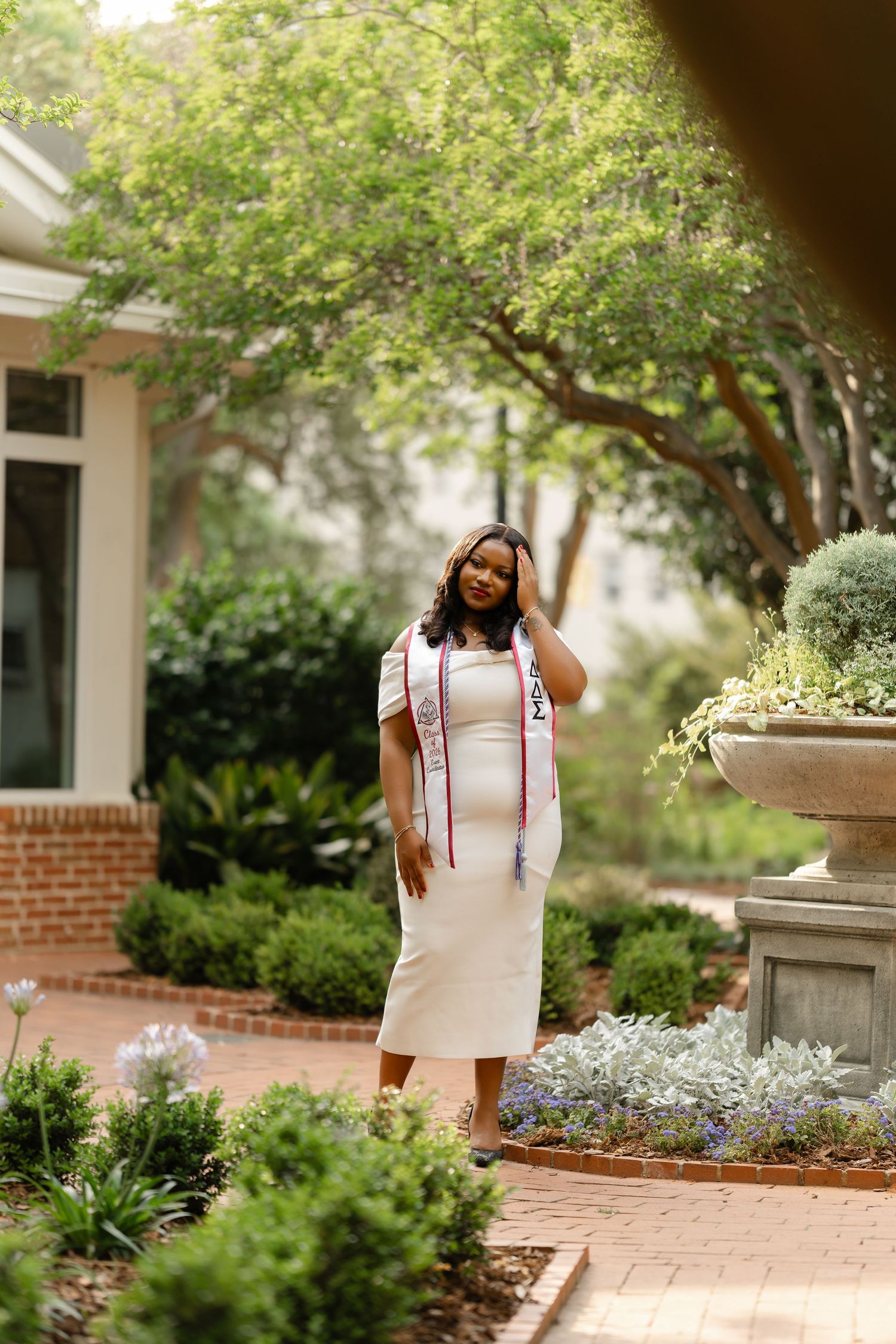 Young woman in white dress and graduation sash poses in a lush garden courtyard during senior graduation photos.
