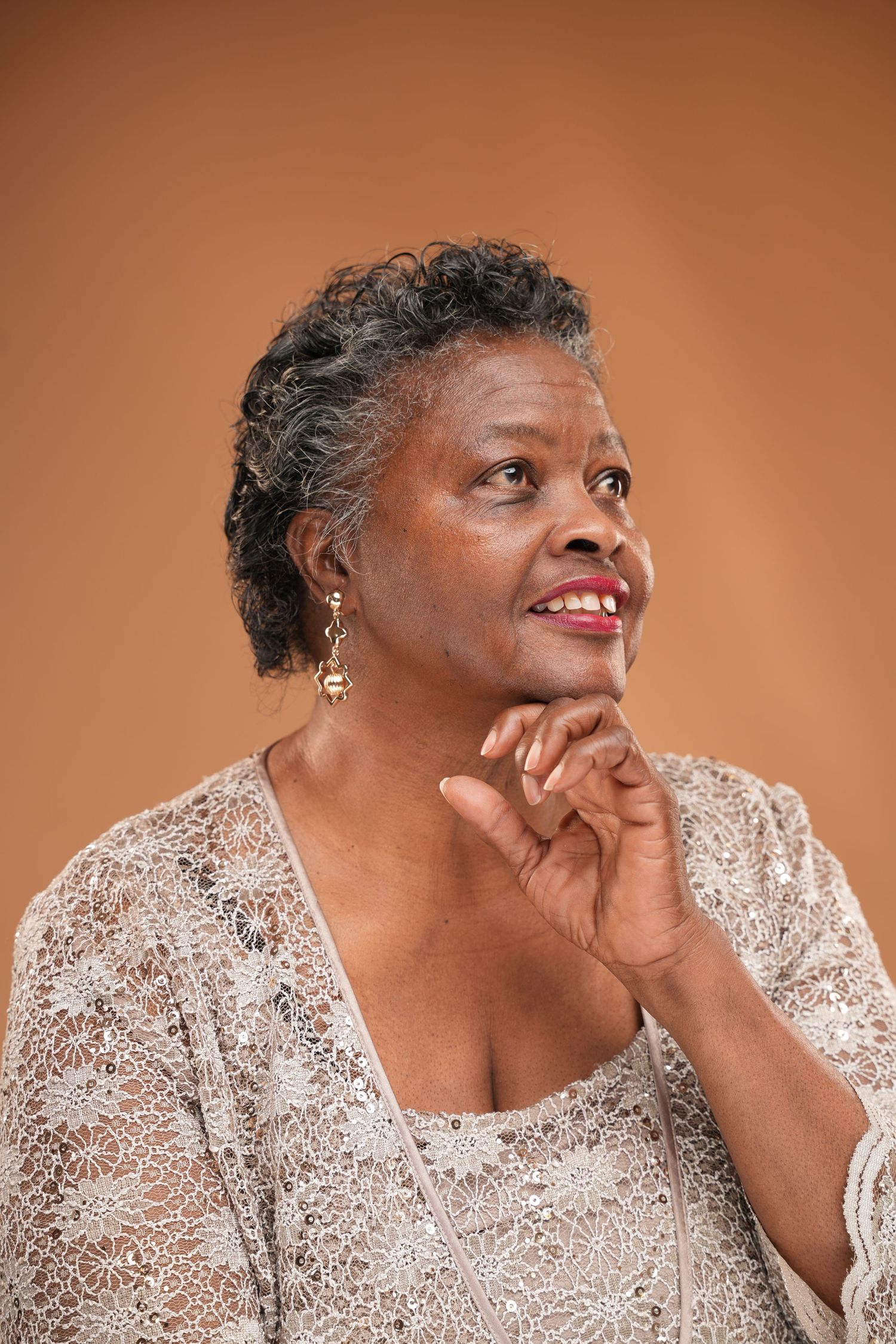 Smiling senior African American woman in lace top, resting chin on hands, looking up thoughtfully on brown background.