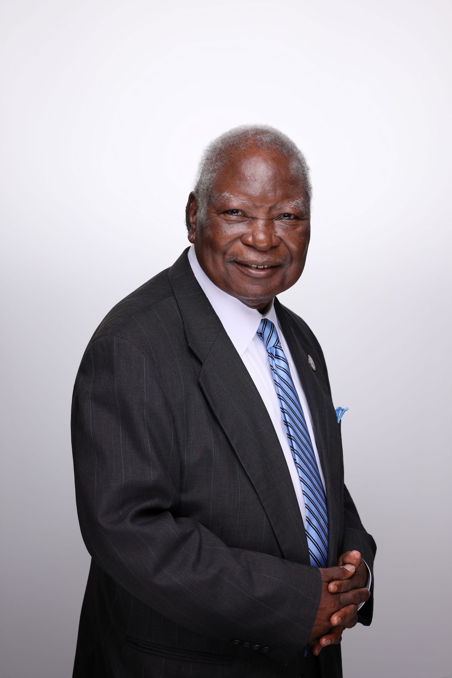 Elderly man in dark suit and blue striped tie smiles warmly in professional portrait photo.