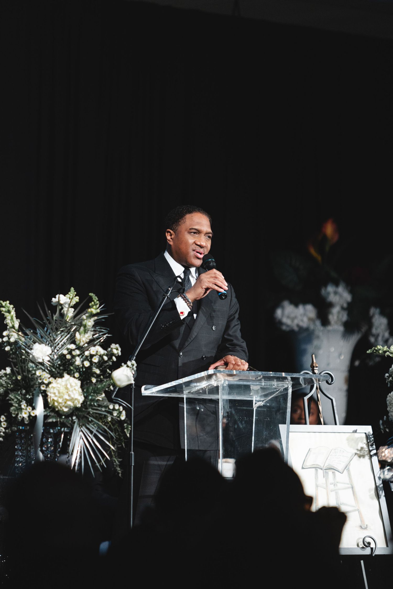 A speaker stands at a clear acrylic podium with white floral arrangements on stage during an evening event.