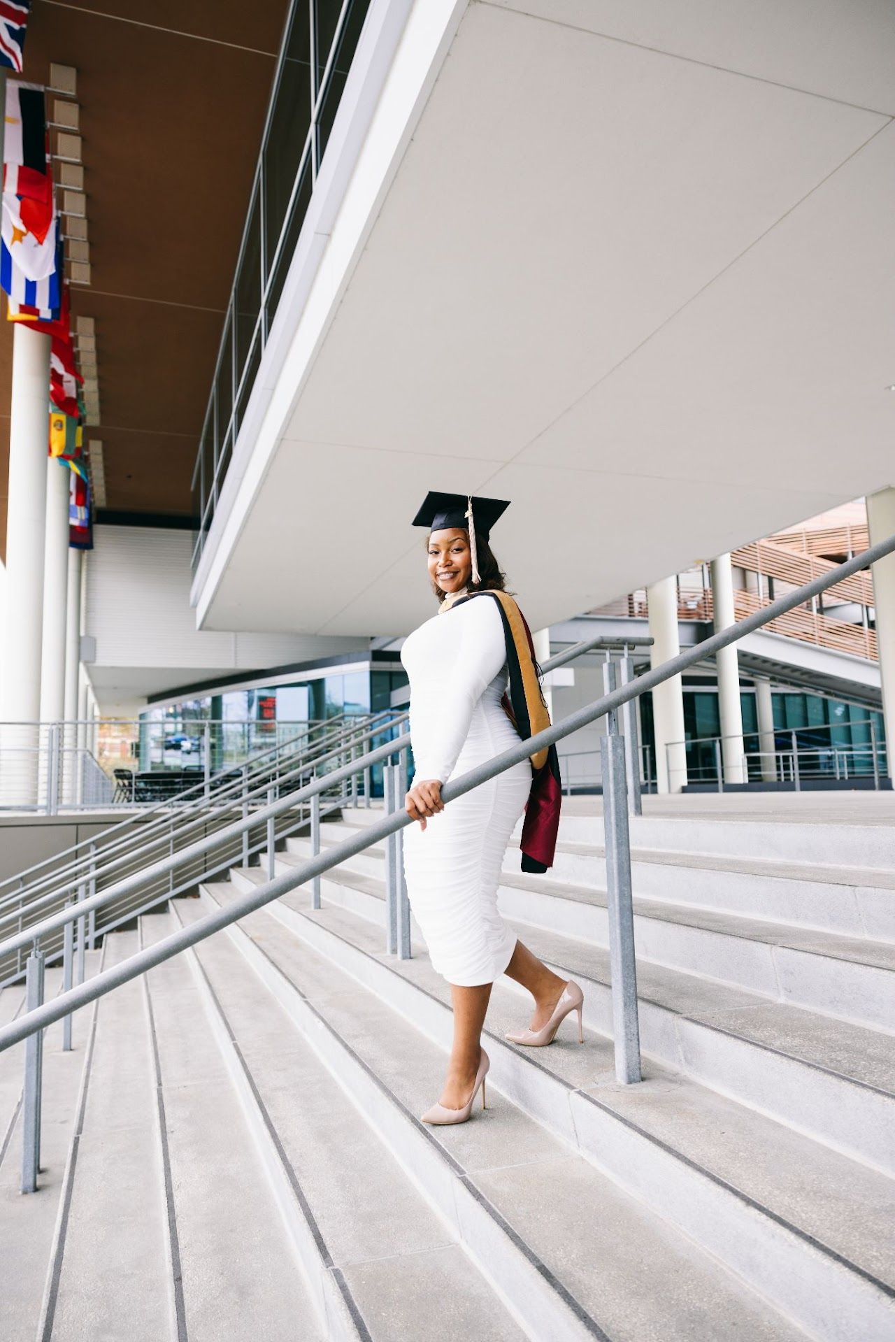 Graduate in white dress and cap ascending concrete steps of modern building with flags.