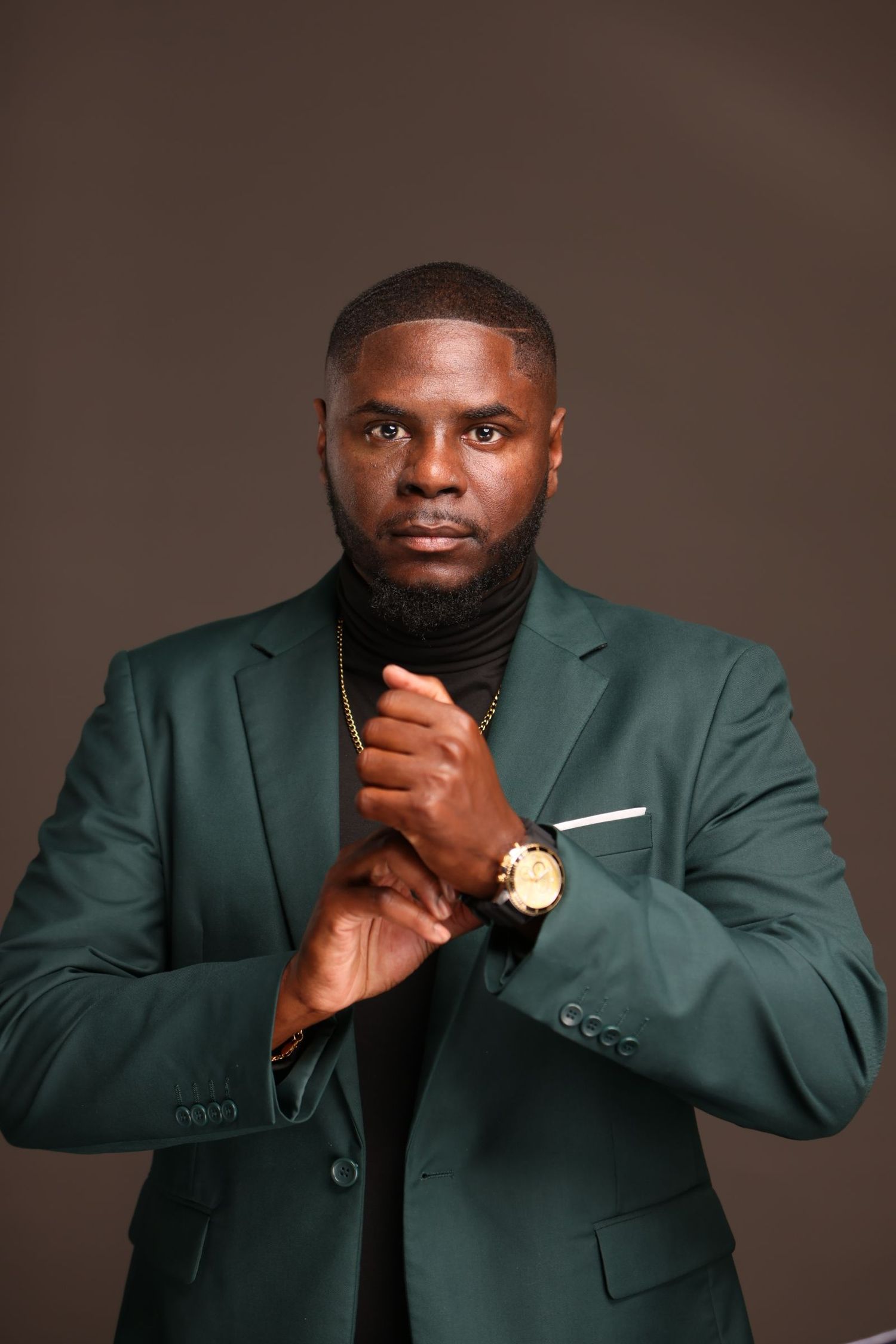 Professional portrait of person in green blazer and black turtleneck with gold watch against dark background.