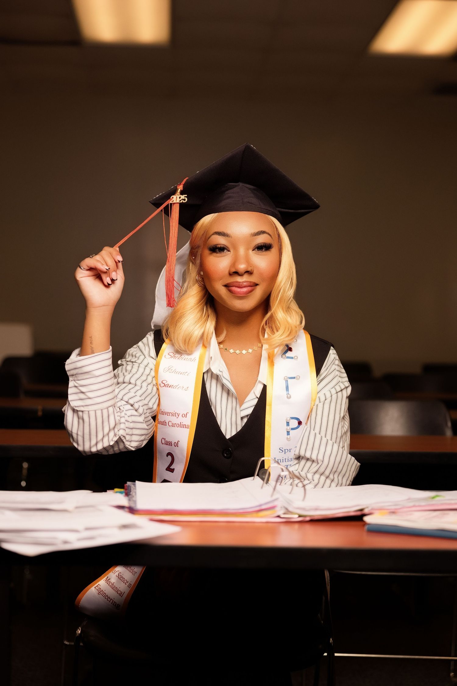 Young woman in graduation cap and sash sits at desk with books, smiling and holding tassel.