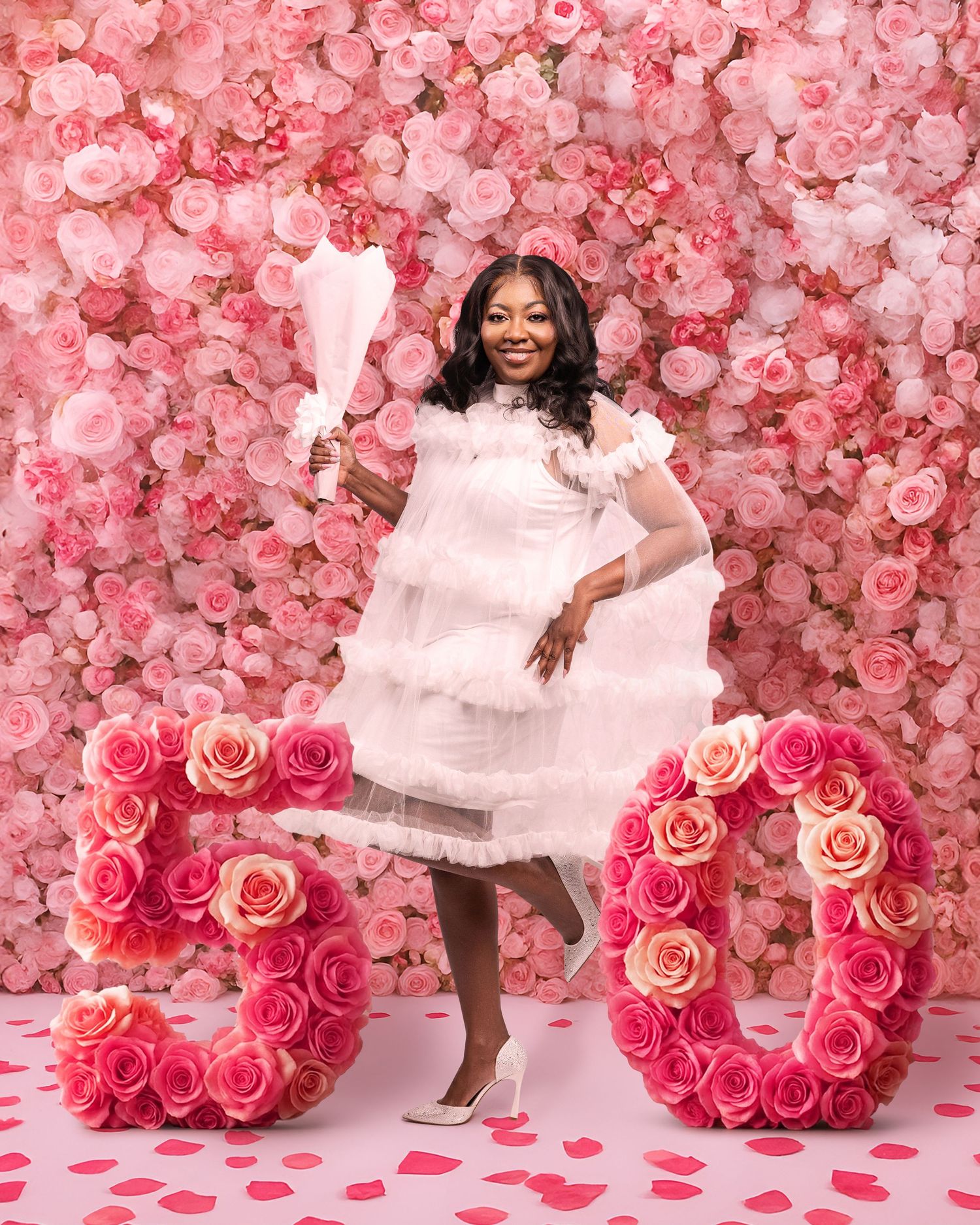 Woman in white dress poses with rose number 50 display against pink floral wall backdrop.