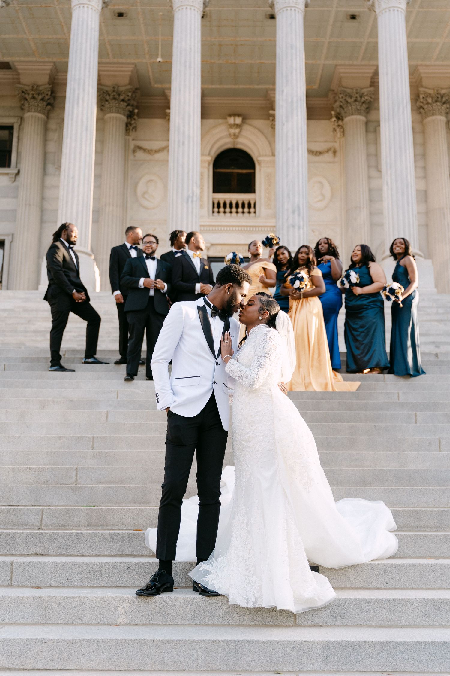 Bride and groom share a kiss on grand stone steps surrounded by wedding party in blue gowns and tuxedos.