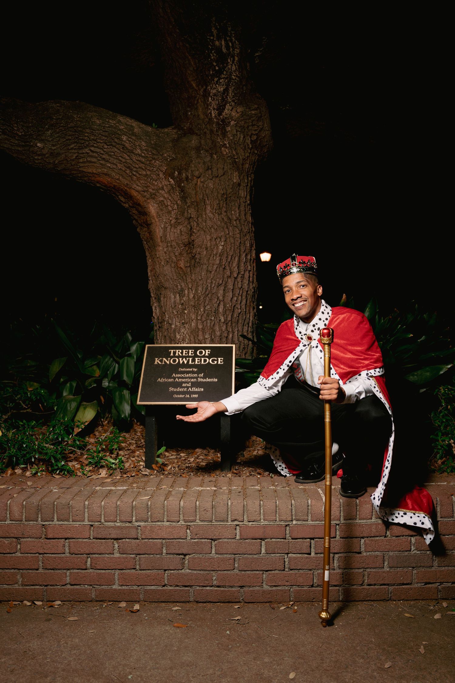 Man in royal king costume poses by Tree of Knowledge plaque at night, holding golden scepter with red cape.