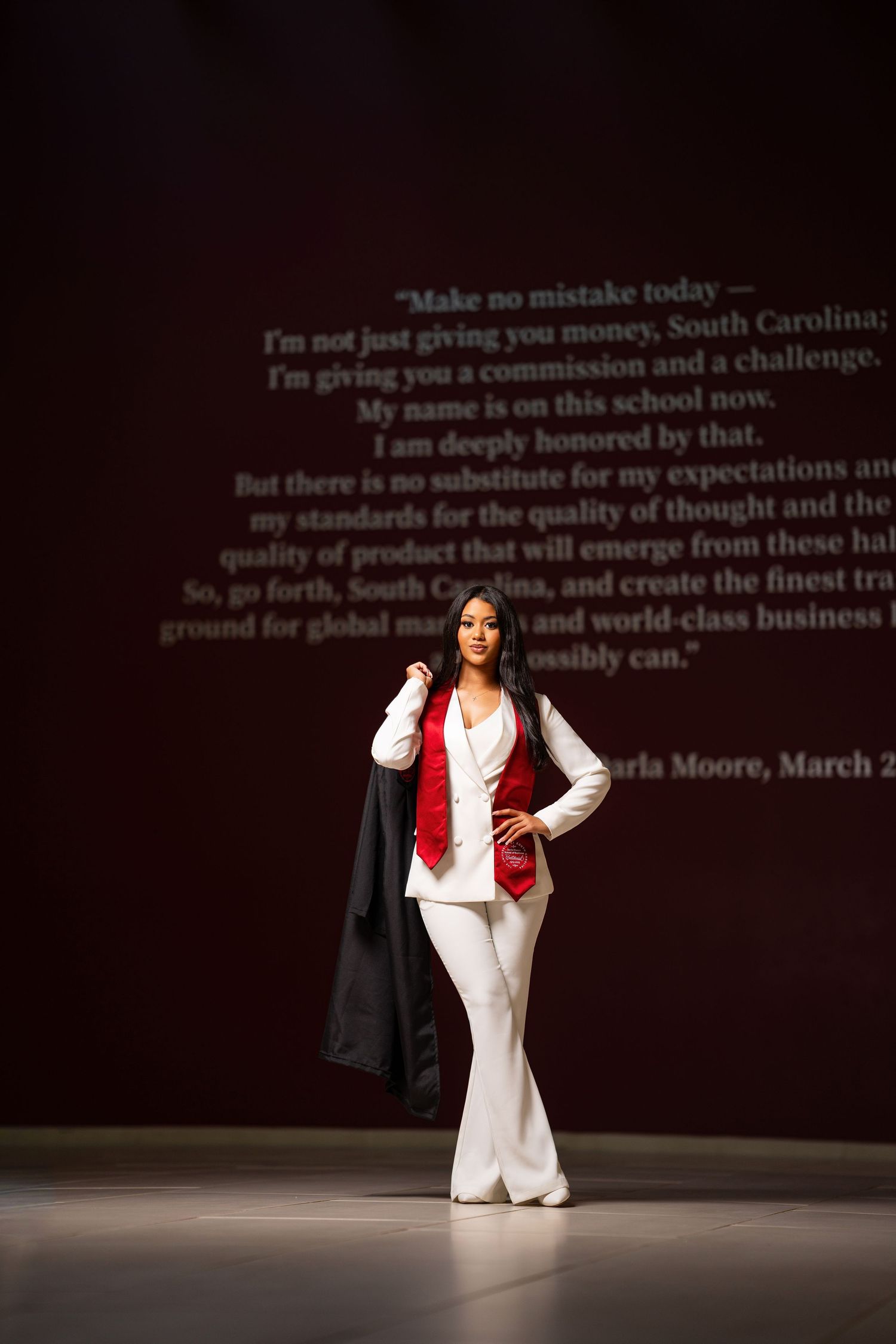 Woman in white suit and red vest holds black jacket on stage before Darla Moore quote projection.