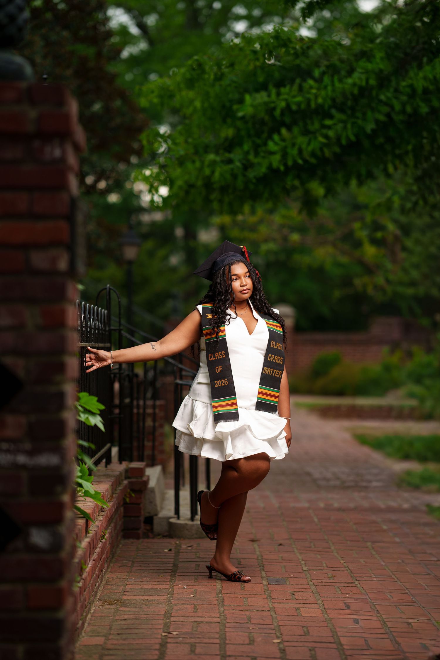 Young woman in white dress and kente cloth graduation stole posing on brick pathway surrounded by lush greenery.