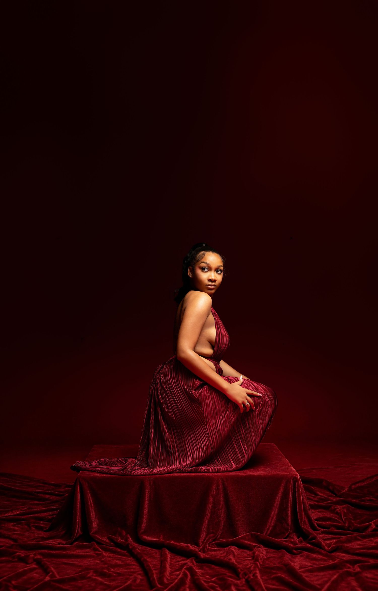 Woman in elegant red gown seated on velvet draped platform against deep crimson studio background.