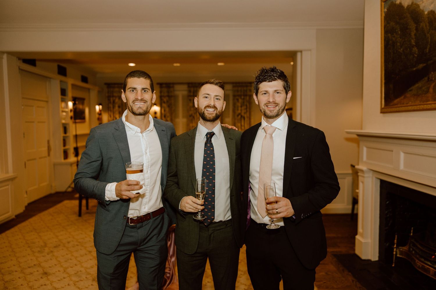 Three people in formal suits holding drinks while standing in an elegant hallway with a fireplace.