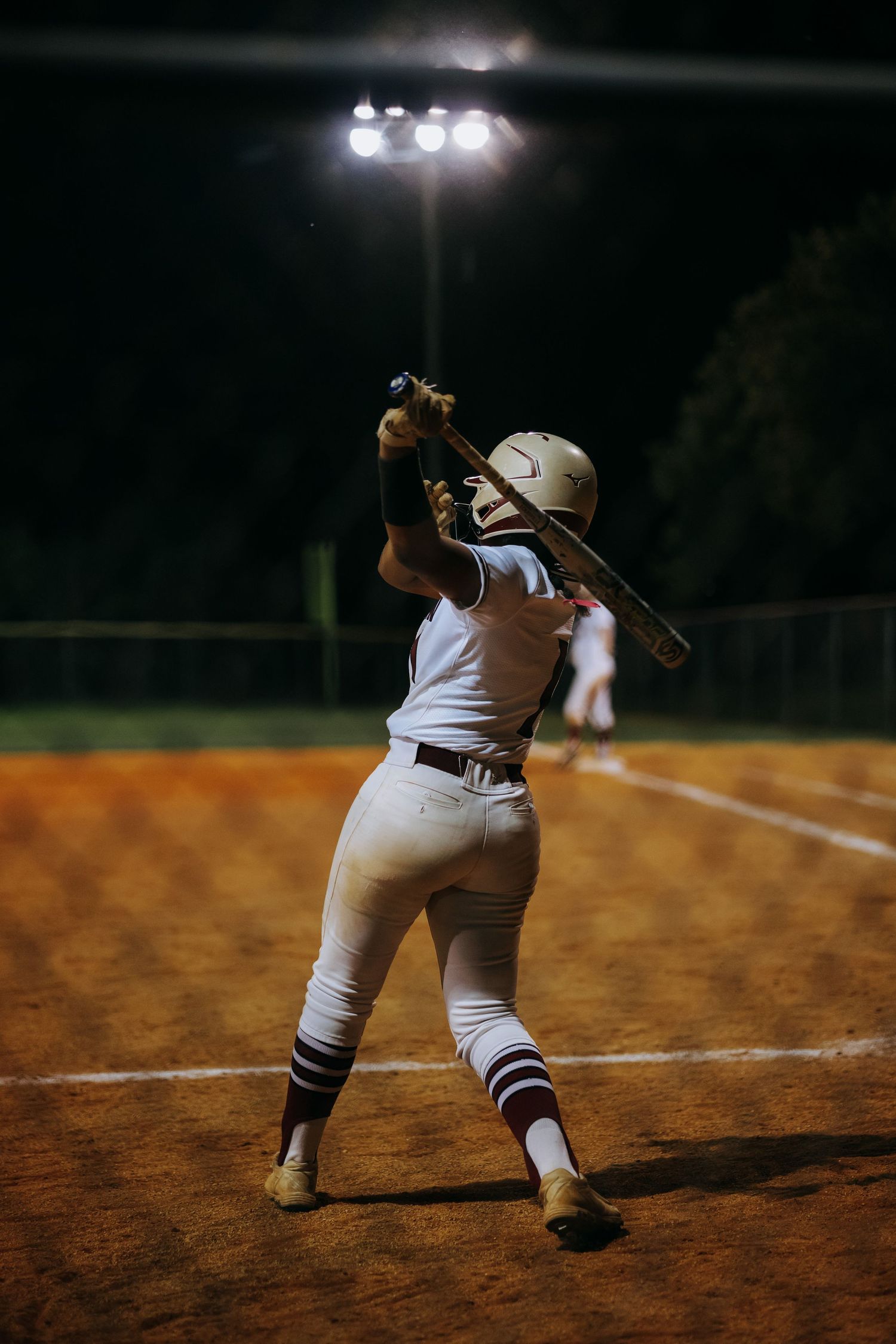 Softball batter in batting stance under bright stadium lights at night.