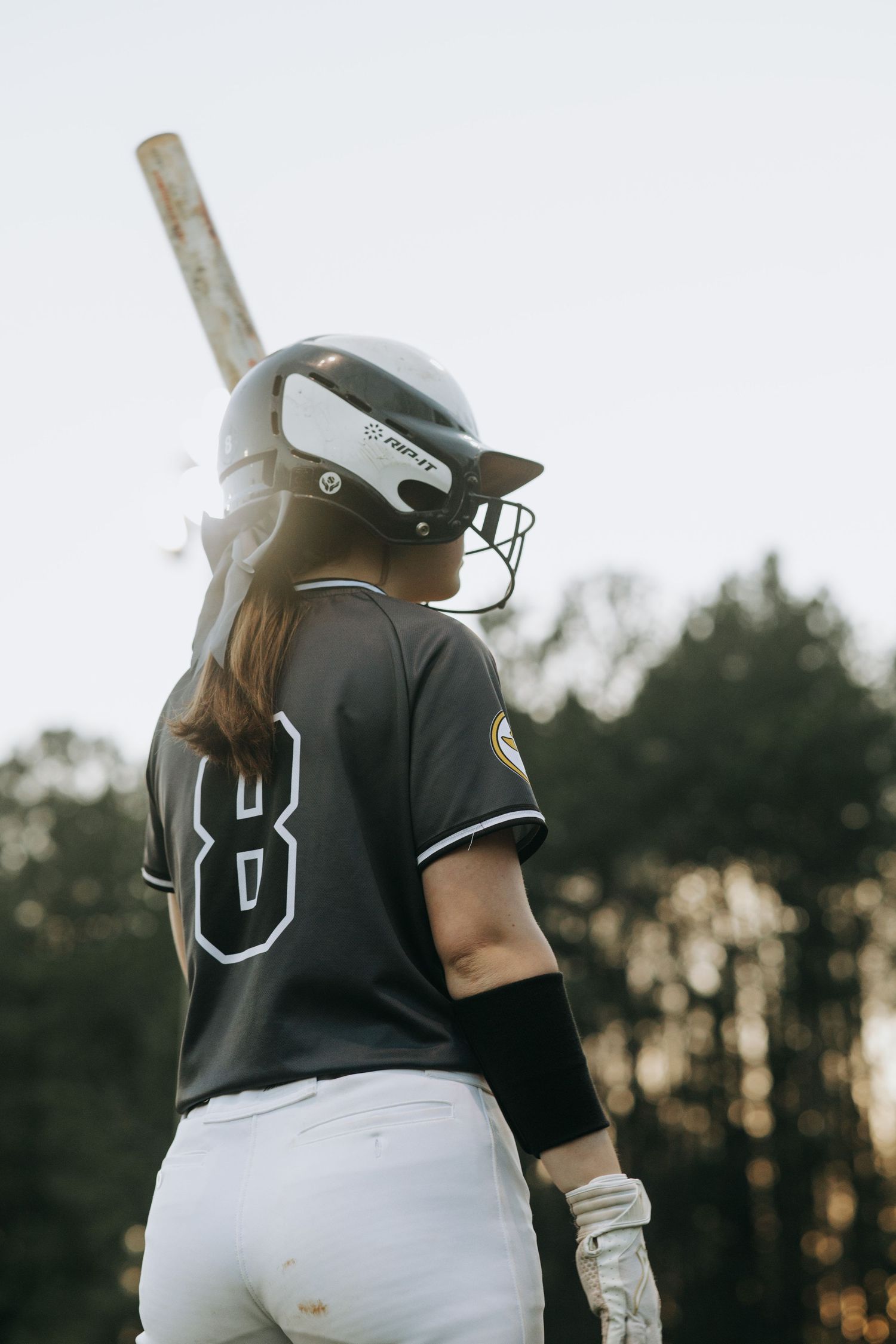 Softball batter in black jersey and white pants prepares to swing at dusk.