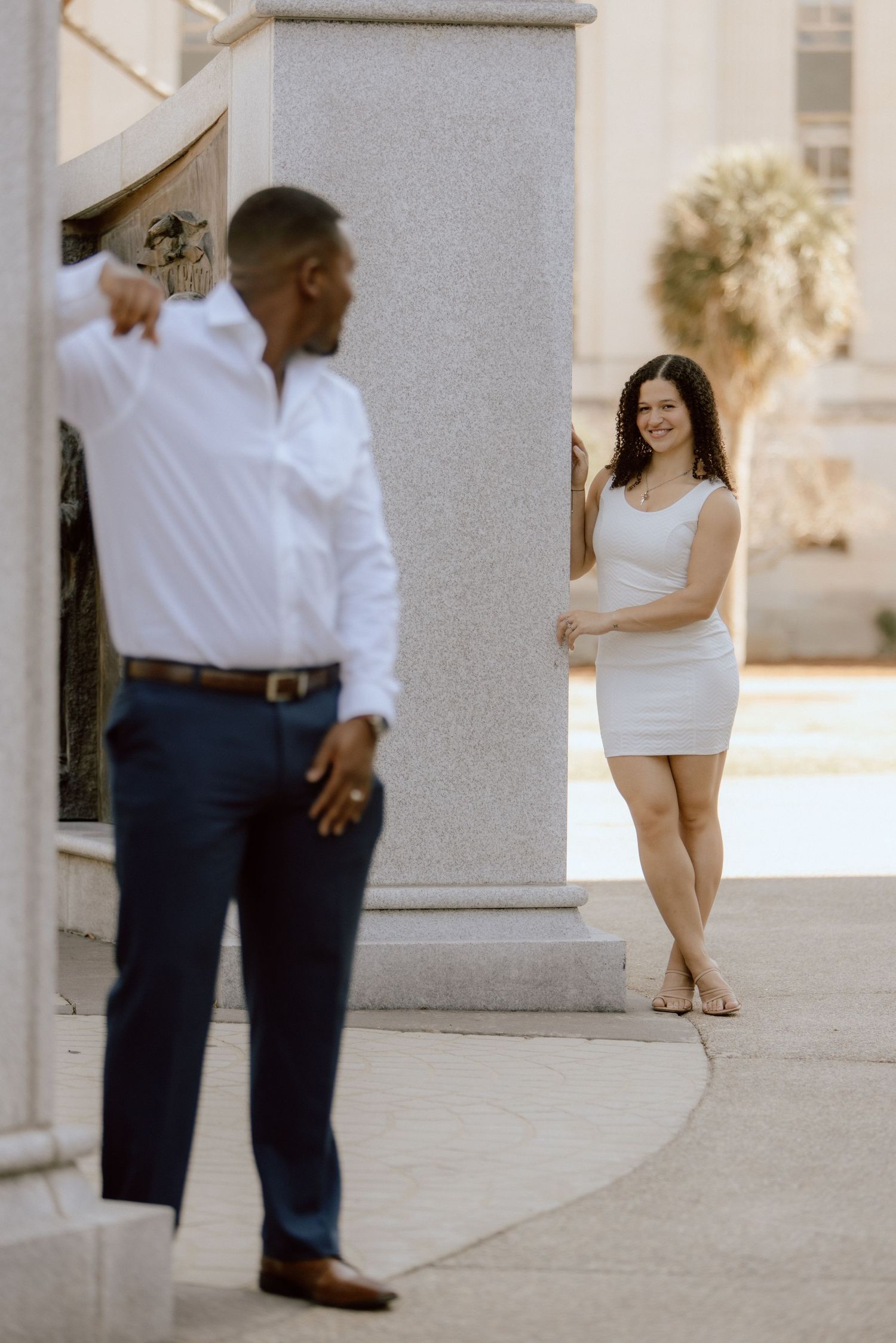 Couple shares playful moment around marble column in formal attire.