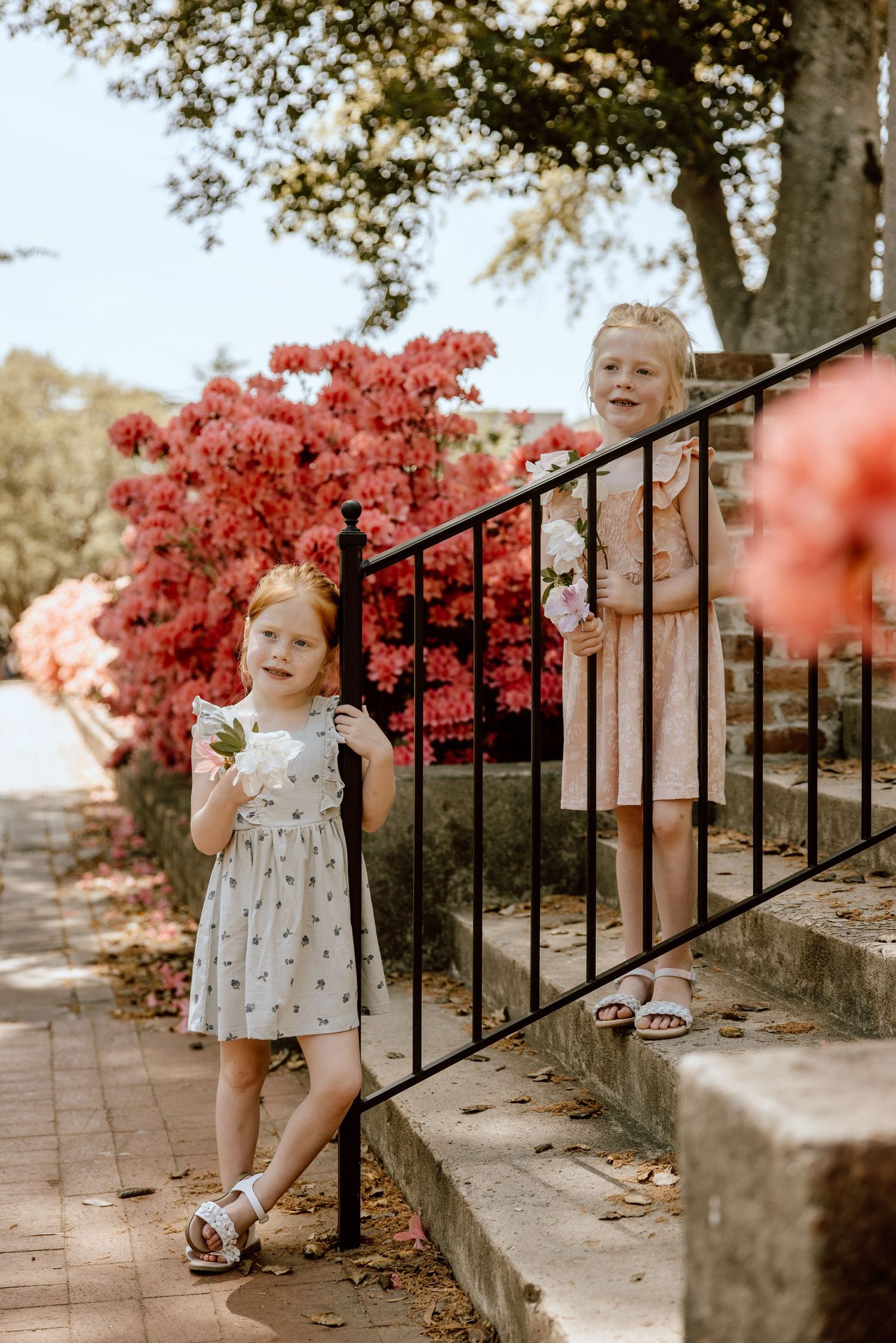 Children in summer dresses playing near red flowers and metal railing outdoors.