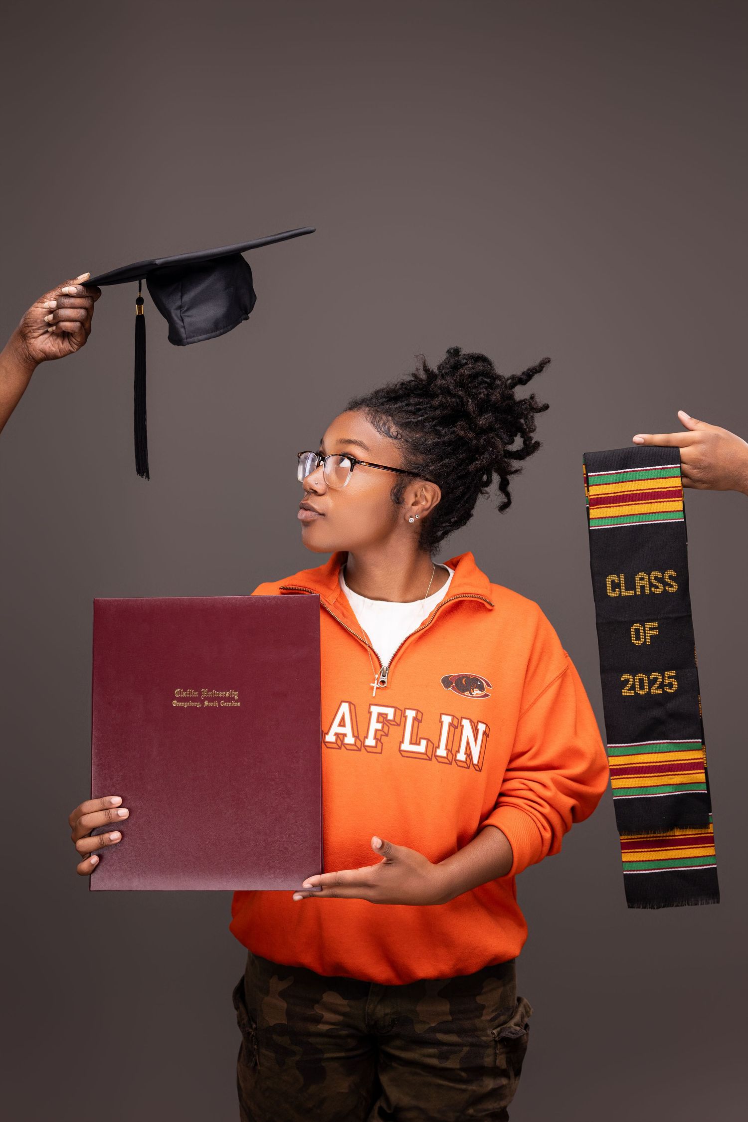 Graduate in orange Claflin sweatshirt displaying diploma and graduation accessories.