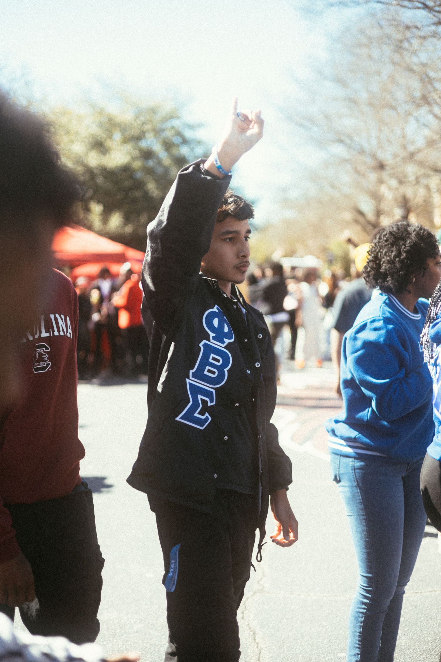 Person wearing a black jacket with Greek letters raises their hand at an outdoor gathering.