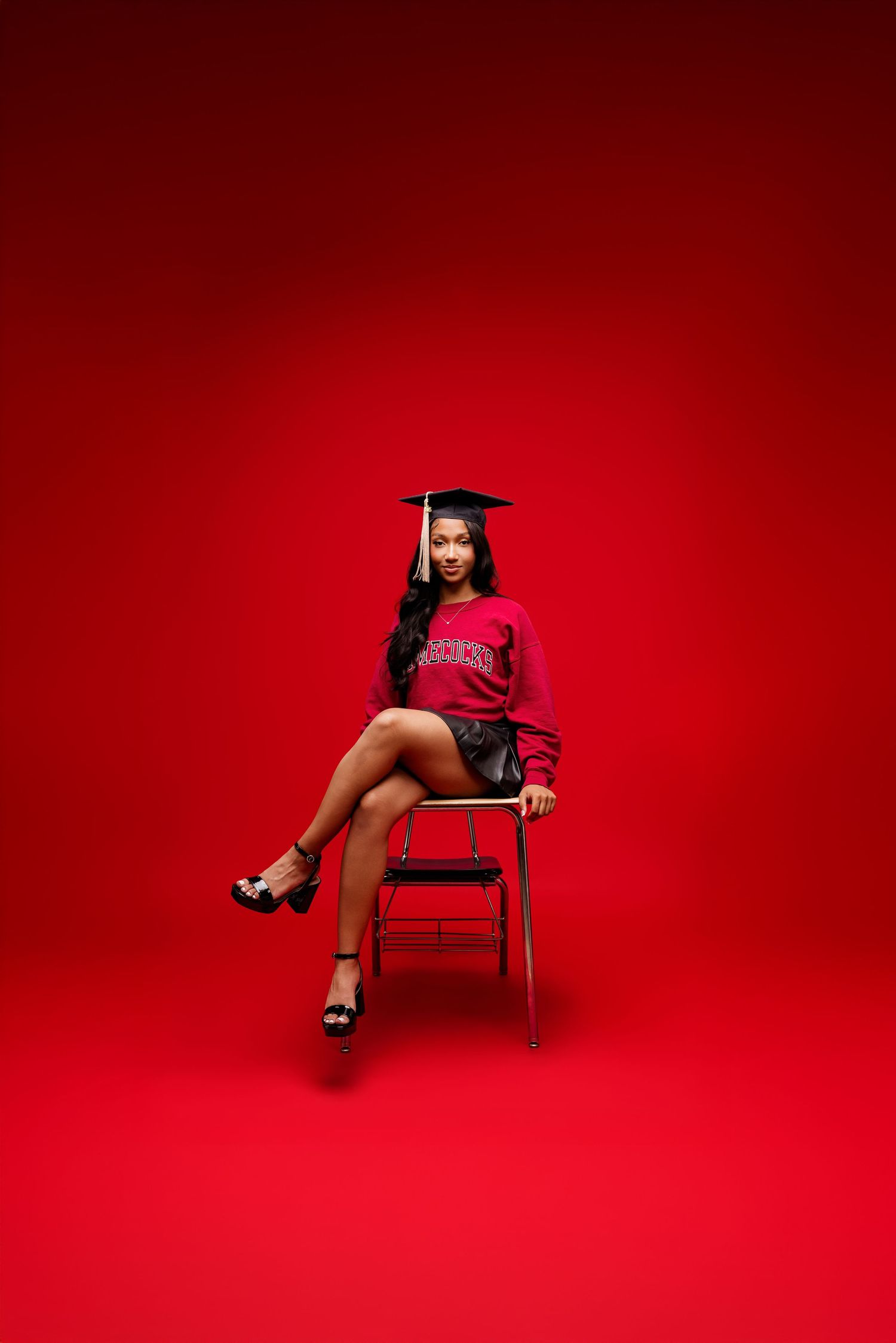 Graduate in pink shirt and mortarboard sits on wooden chair against vibrant red backdrop in studio setting.