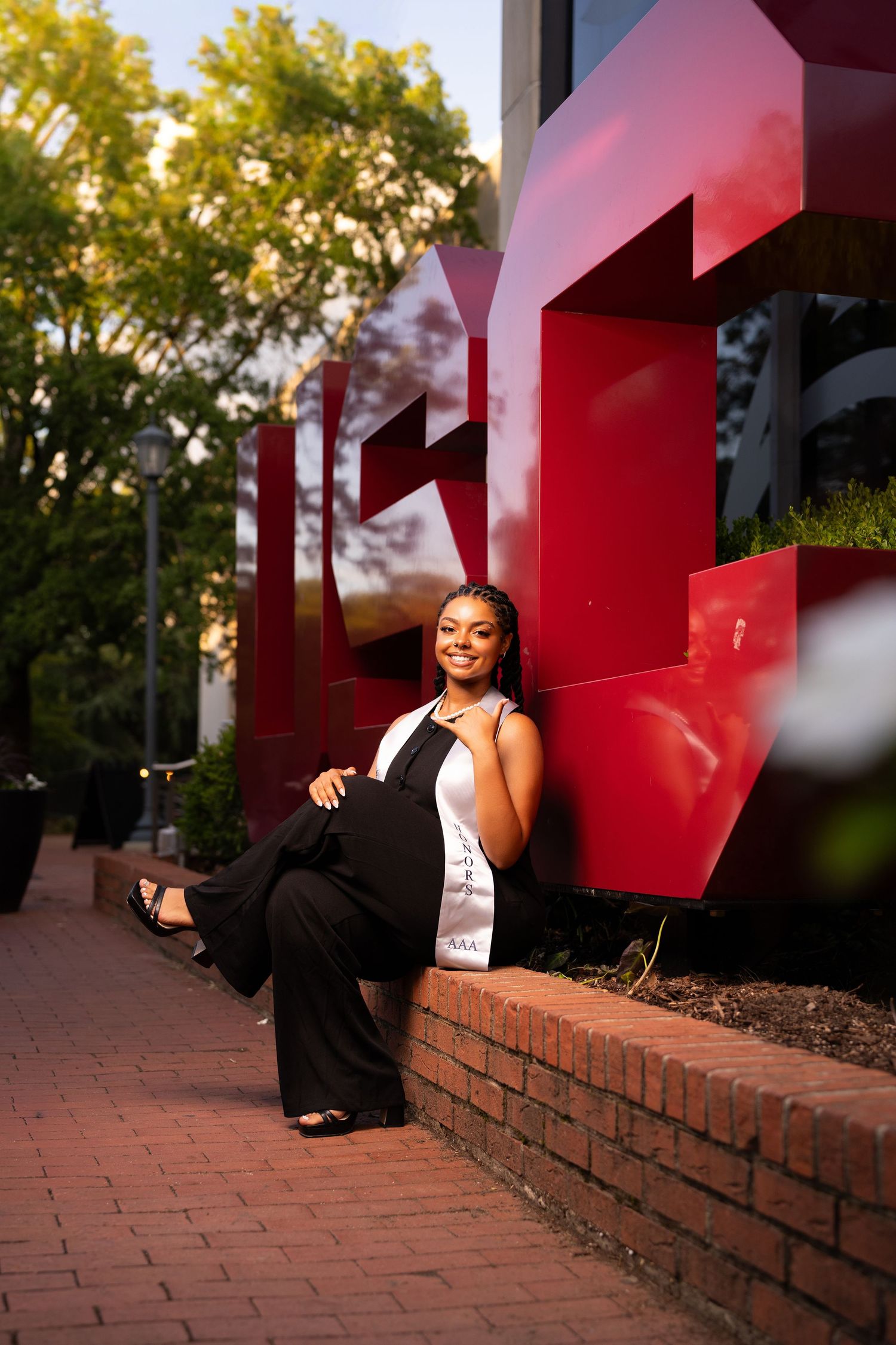 A smiling young woman in black jumpsuit with sash poses by red IU sculpture on brick pathway at golden hour.