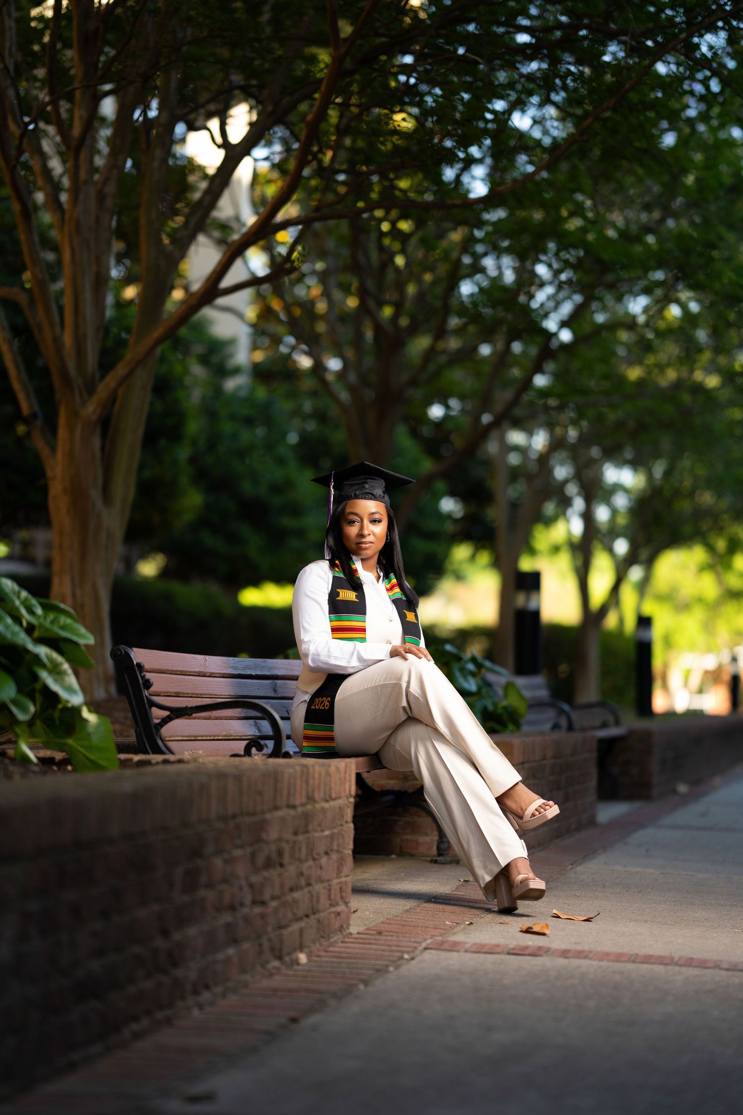 A young Black woman in graduation cap and kente stole sits on a brick bench outdoors among lush green trees.