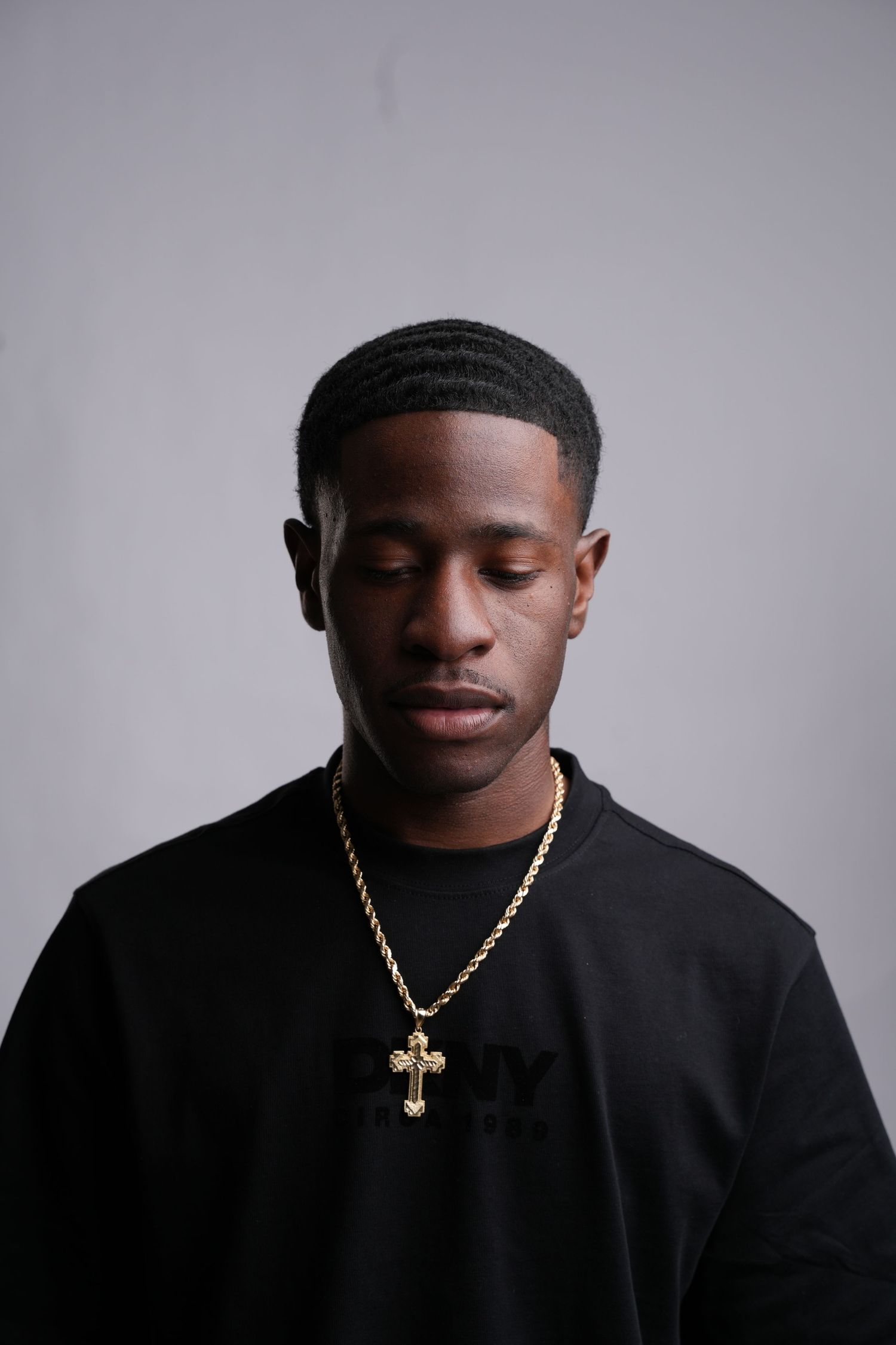 Young Black man in black sweatshirt wearing gold cross necklace, eyes closed, posing against gray background.