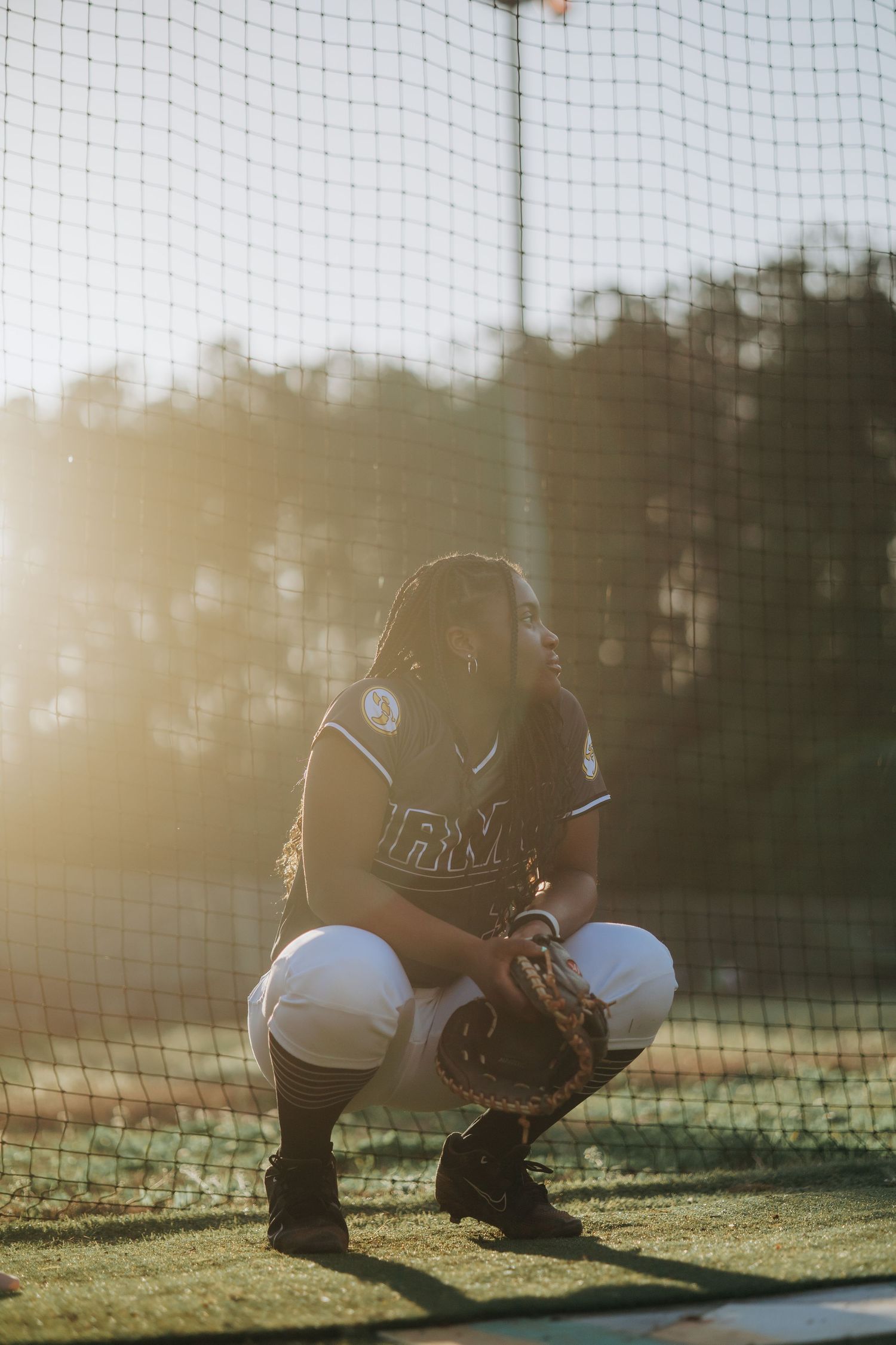 Softball catcher crouches behind home plate netting during golden hour.