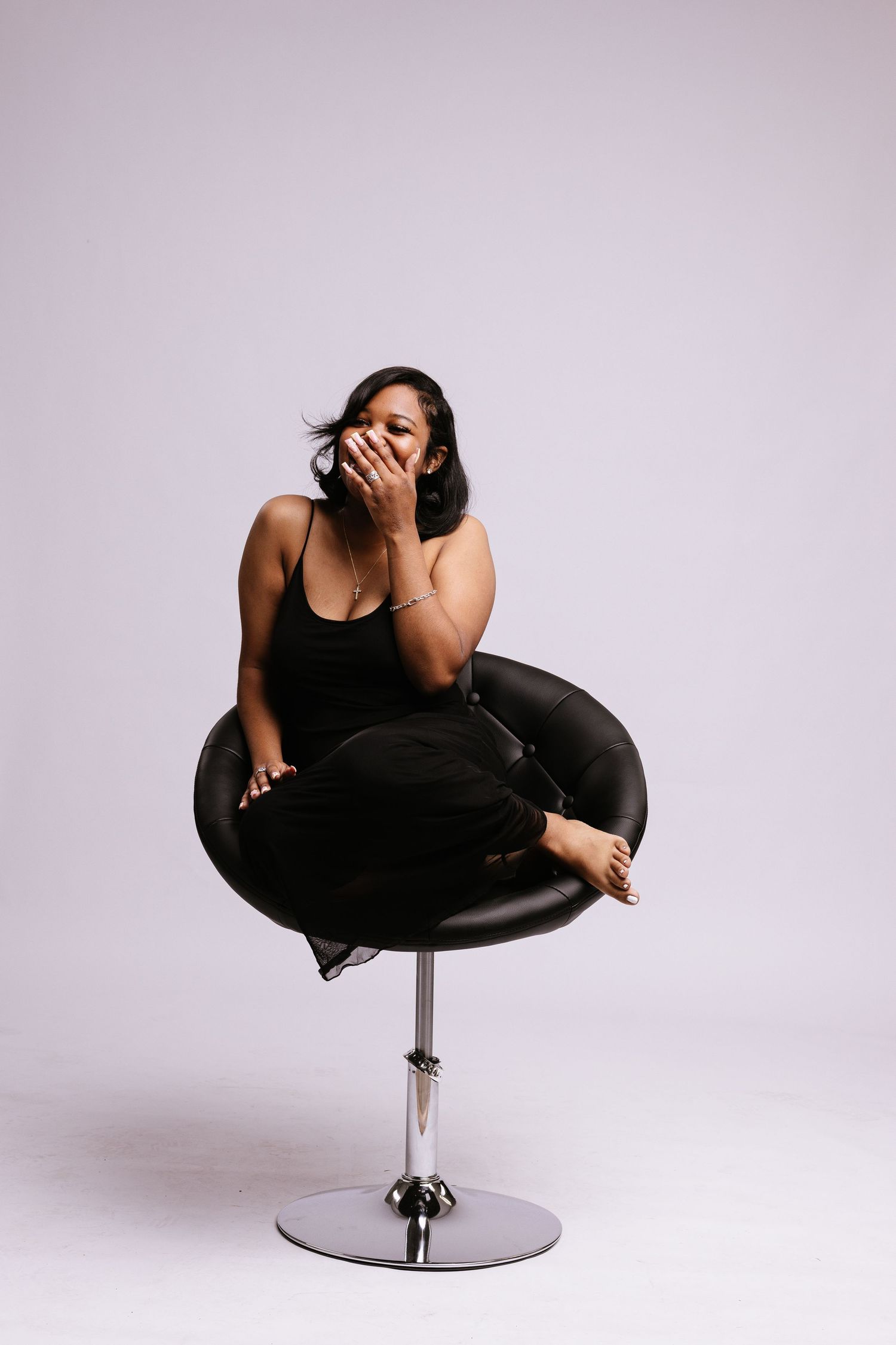 Woman in black outfit relaxes in modern swivel chair against light background.