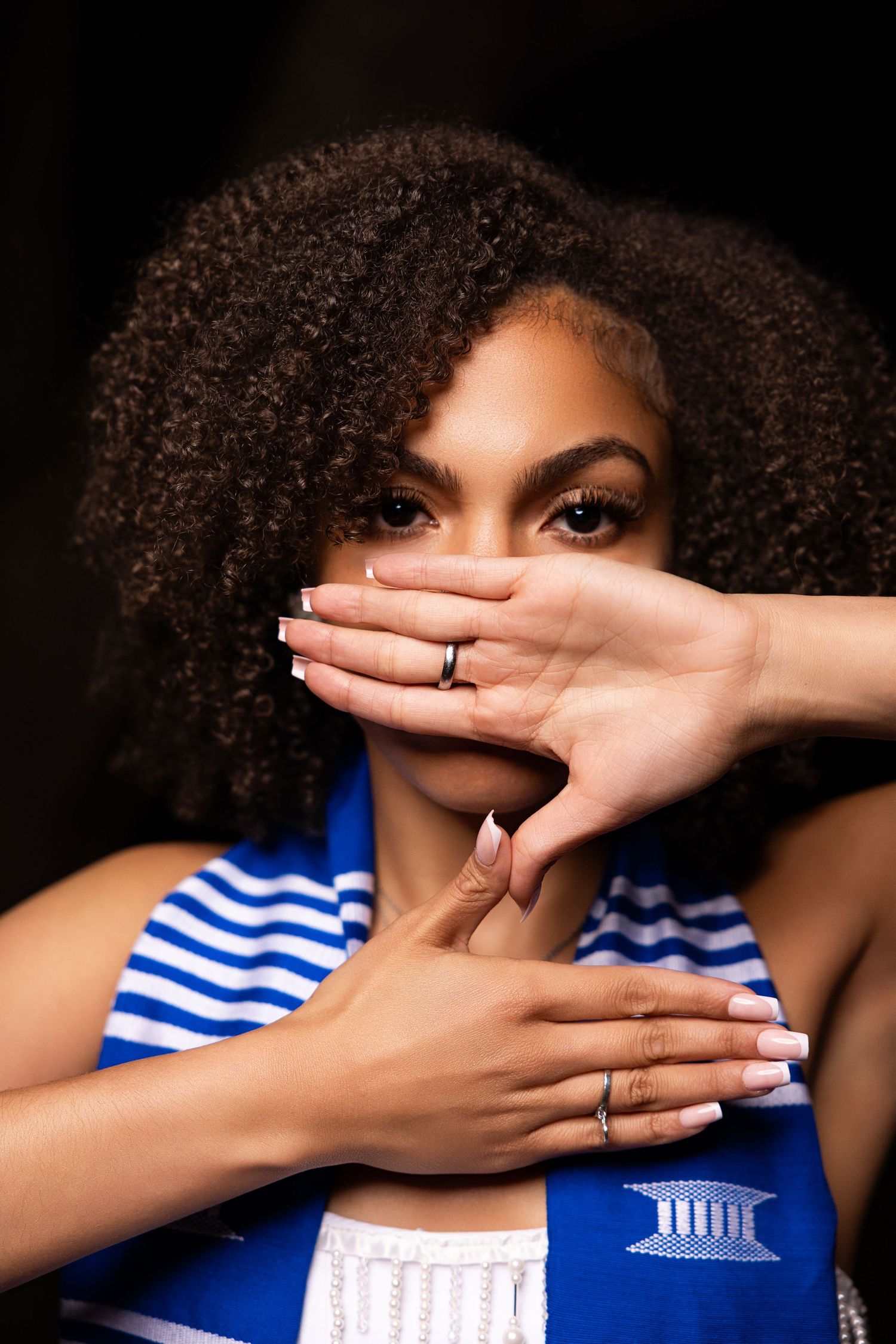 Graduate in blue and white stole shows engagement ring in artistic portrait.
