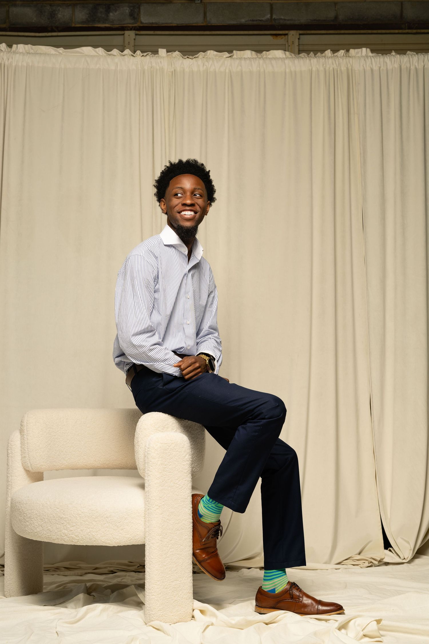 A person wearing a light blue button-down shirt and jeans sits casually on a modern white chair against a curtained backdrop.