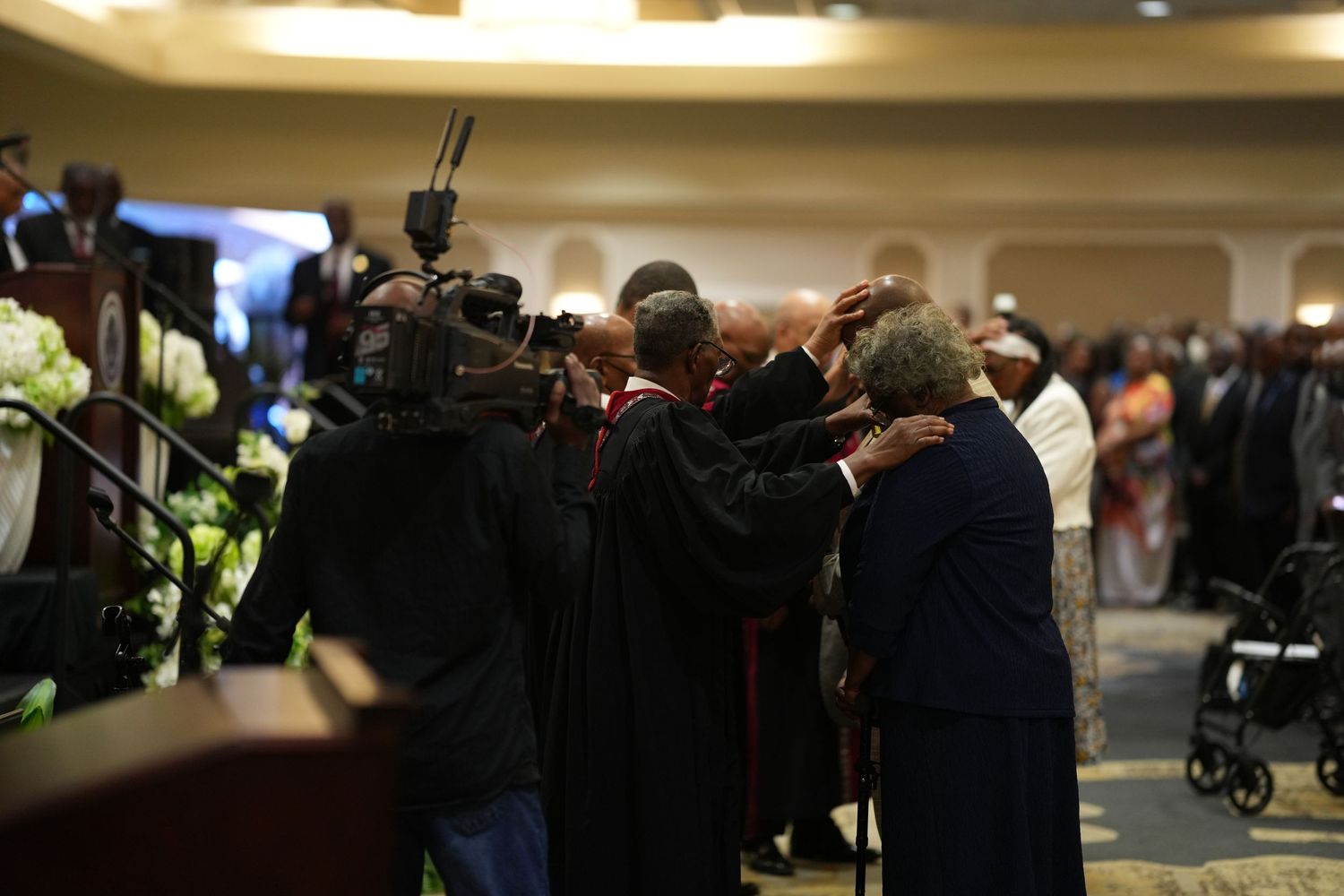 Clergy members in robes gather and pray together at a formal memorial service with camera crews filming.